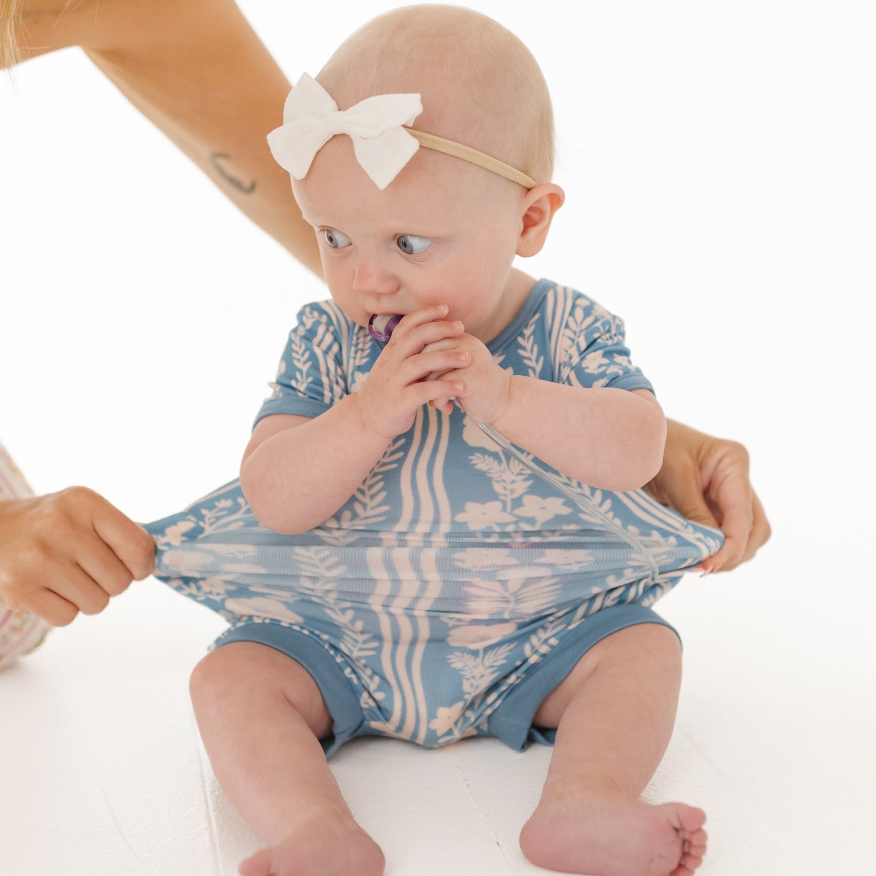 Baby in a blue and white patterned onesie sitting on a white surface with a person holding it.