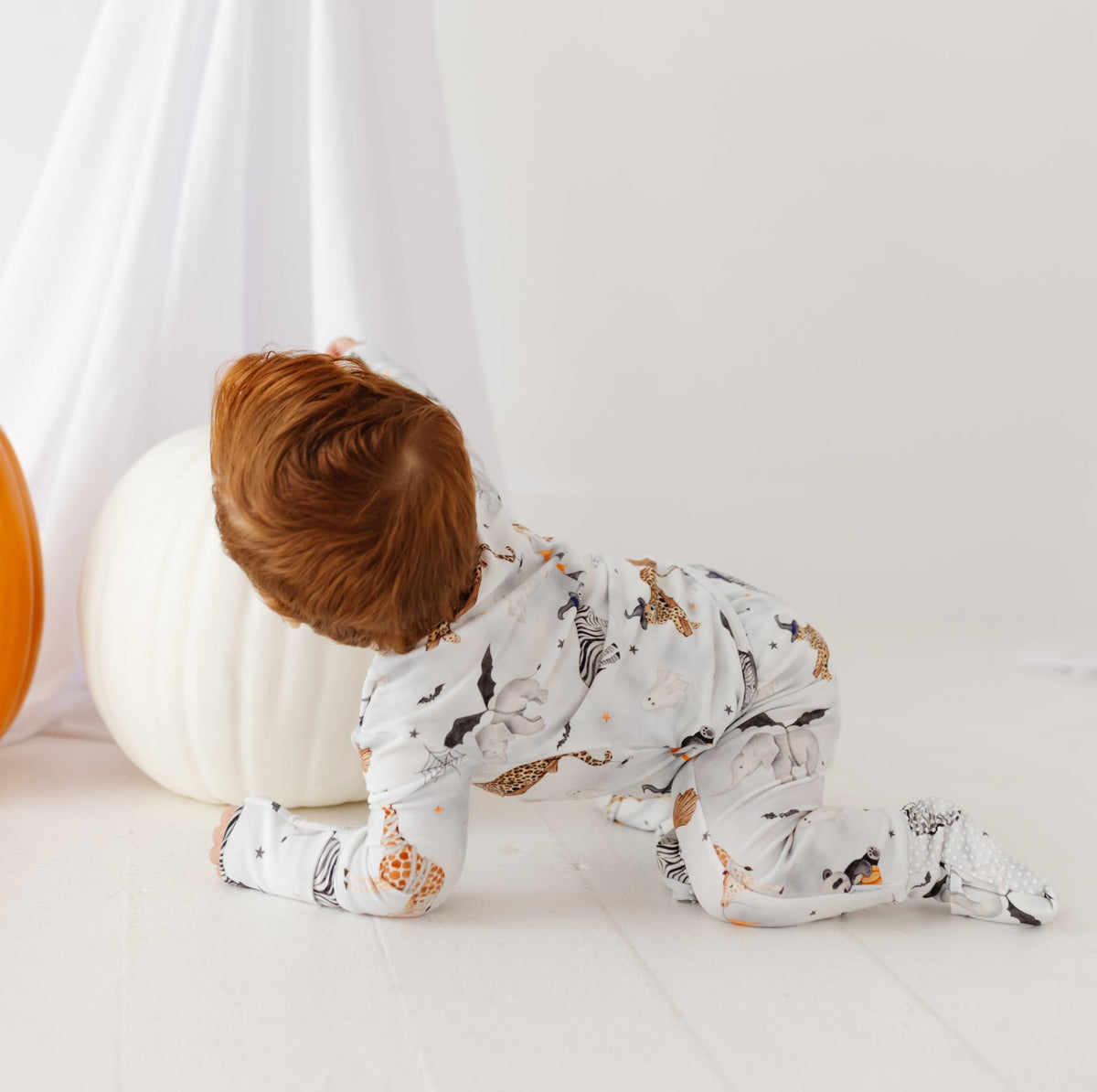 A baby crawls on a white floor with pumpkins, wearing a grey romper with a Halloween-themed print.