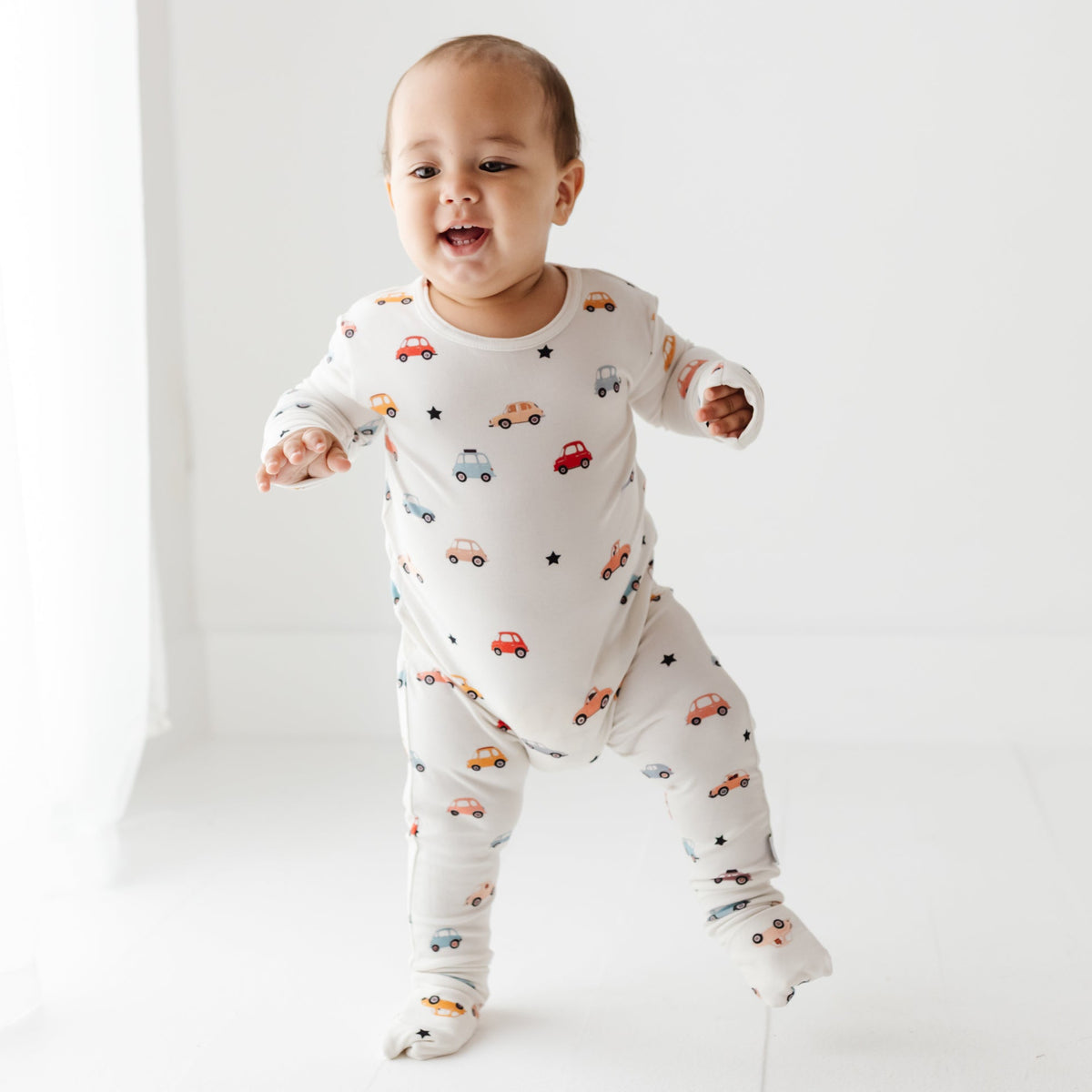 A smiling baby walks on a white floor, wearing a white romper with a car-themed print.