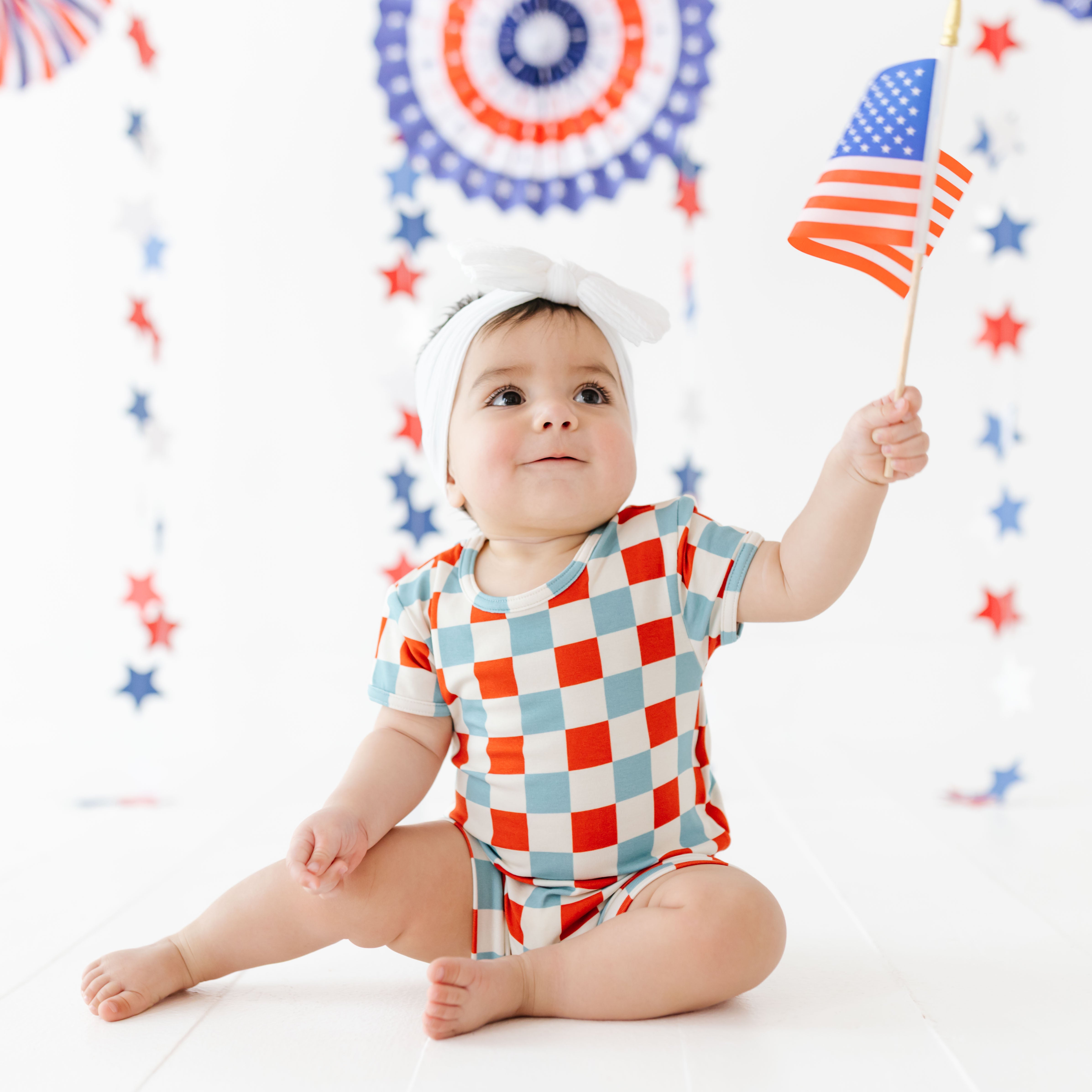 Baby in a checkered outfit holding an American flag with a festive background