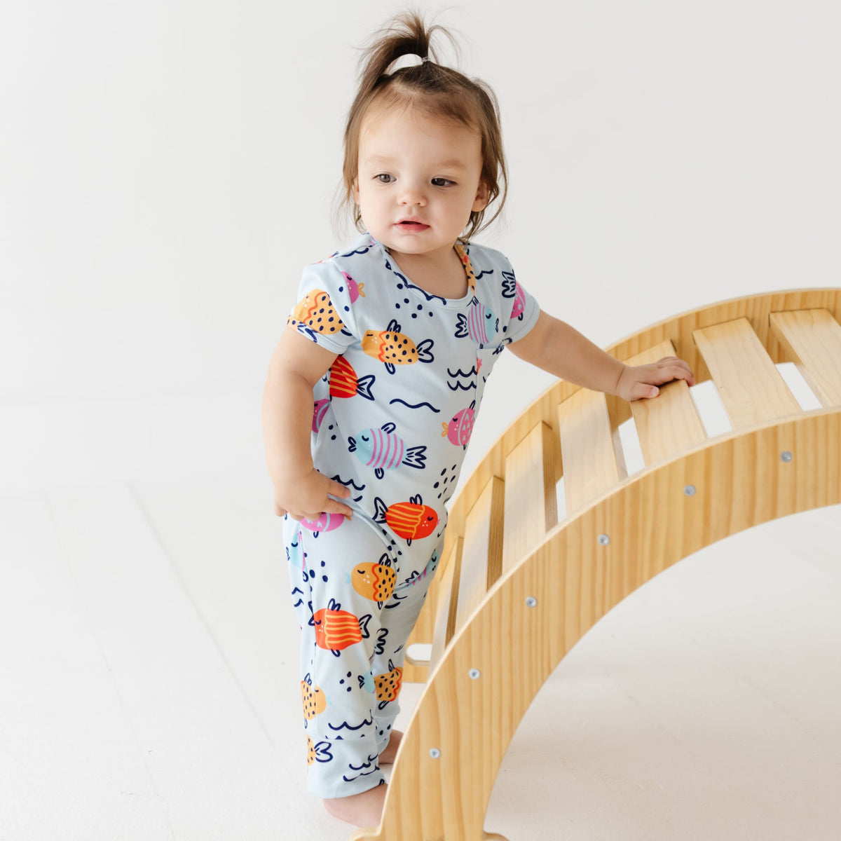 A baby stands against a wooden arch on a white floor. She is wearing a light blue romped with a fish-themed print. 