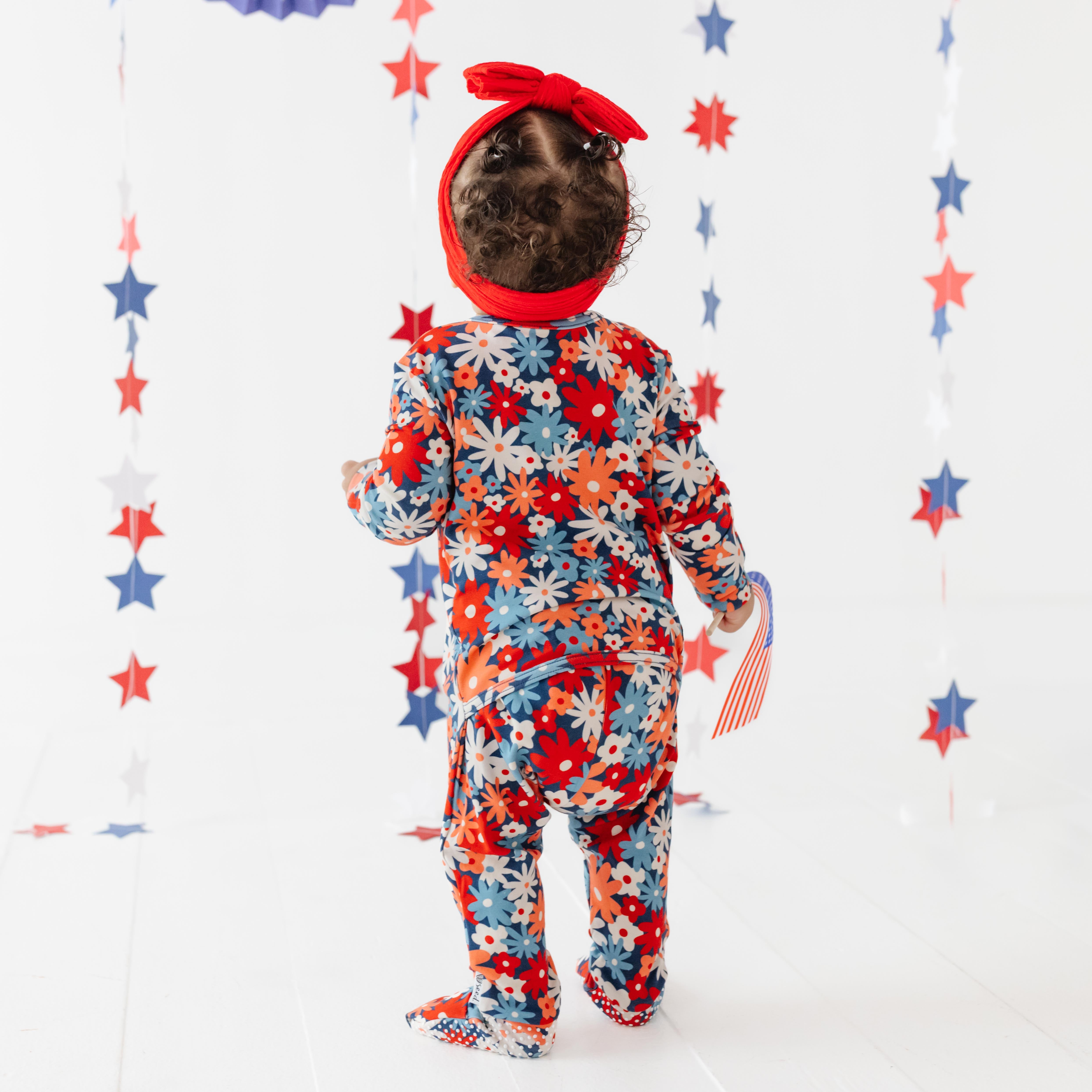 Child wearing a colorful floral onesie with a red bandana, standing against a white background with star decorations.