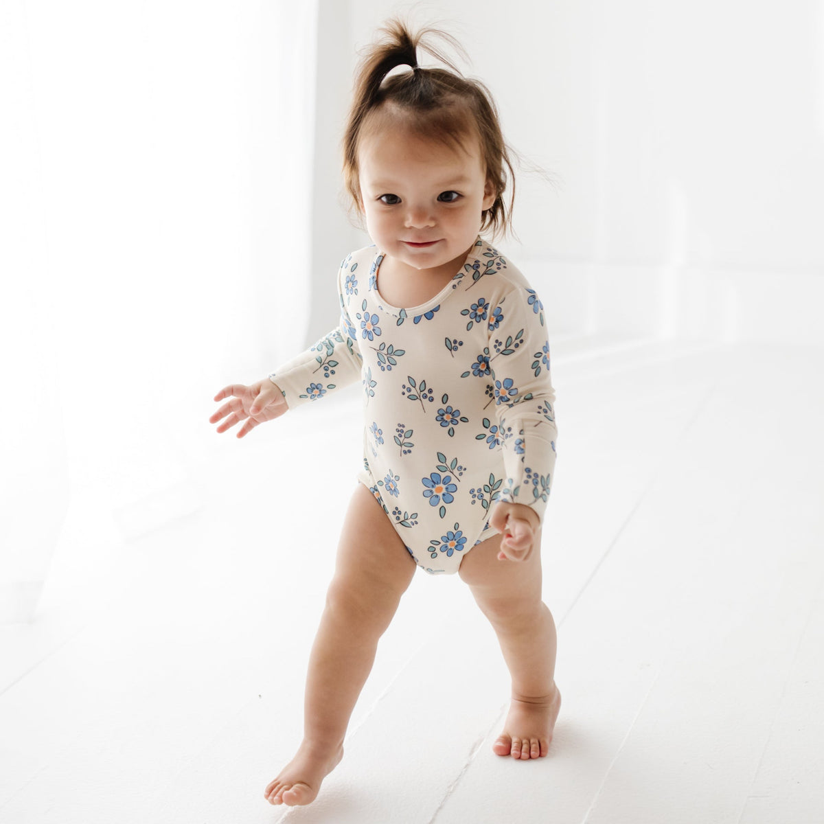 A baby stands on a white floor wearing a cream onesie with blue flowers on it. 