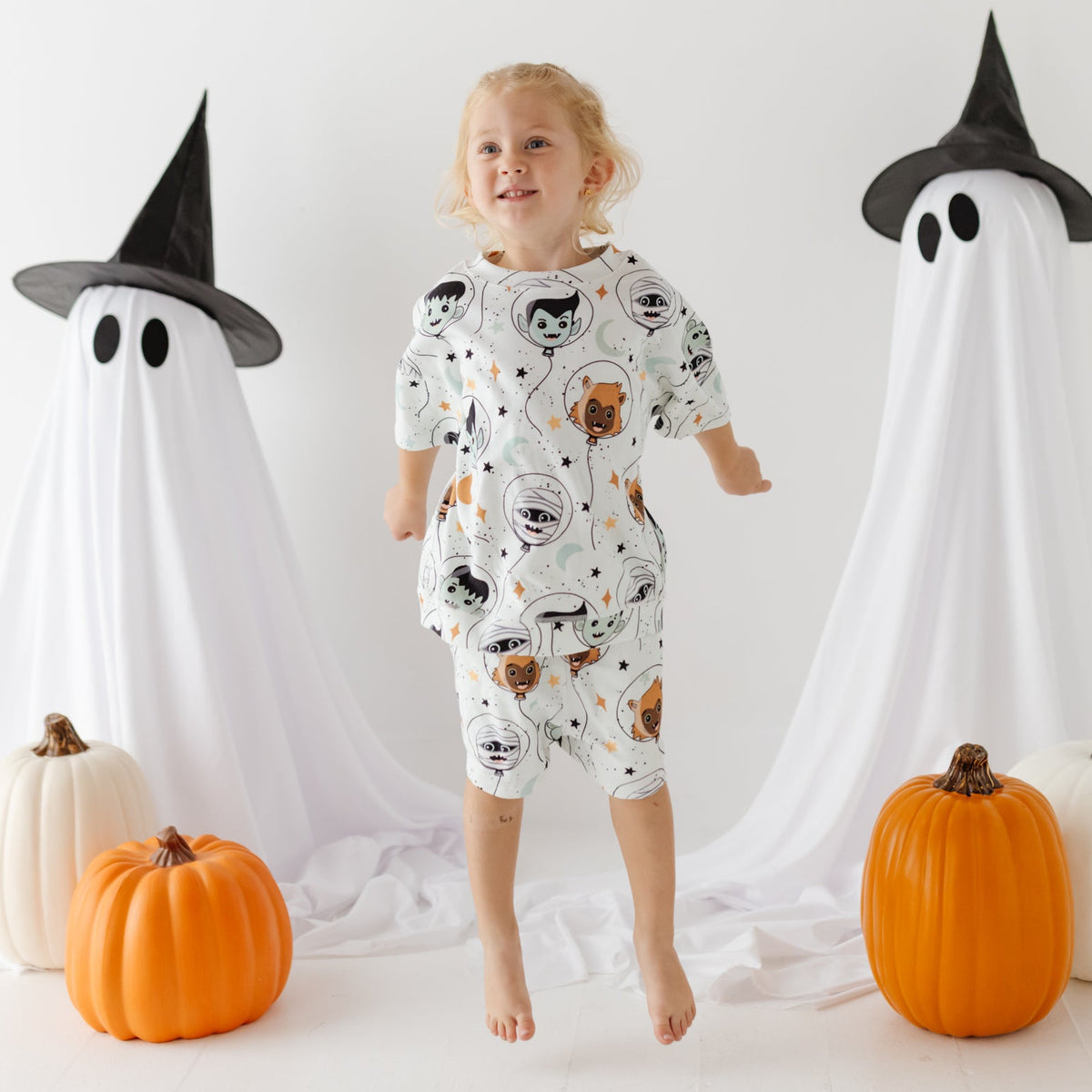 A smiling girl stand on white floor with ghost and pumpkins in the background. She is wearing a white sweatsuit set with a Halloween-themed design. 
