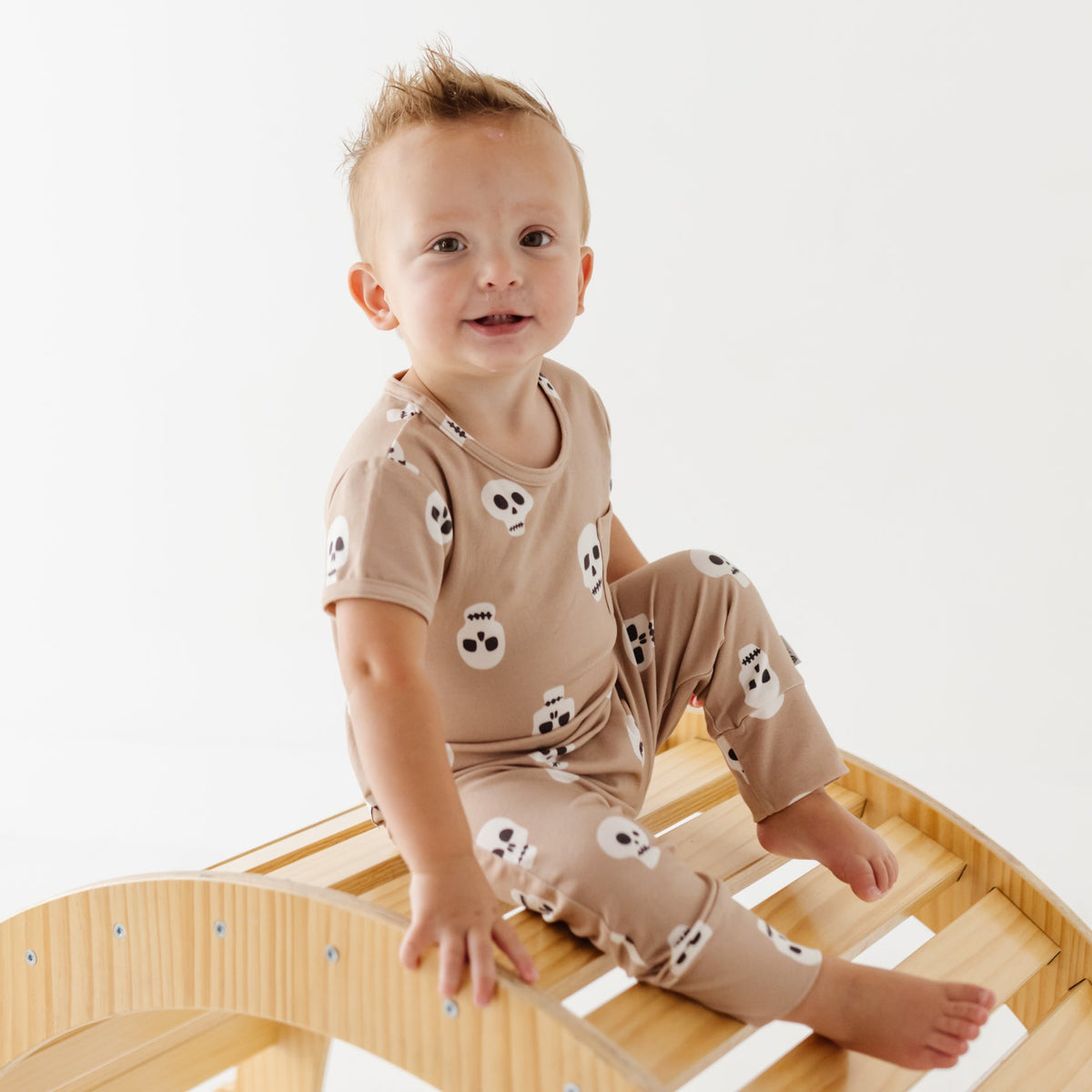 A smiling kid sits on a wooden arch wearing a hand short-sleeved romper with skulls on it. 