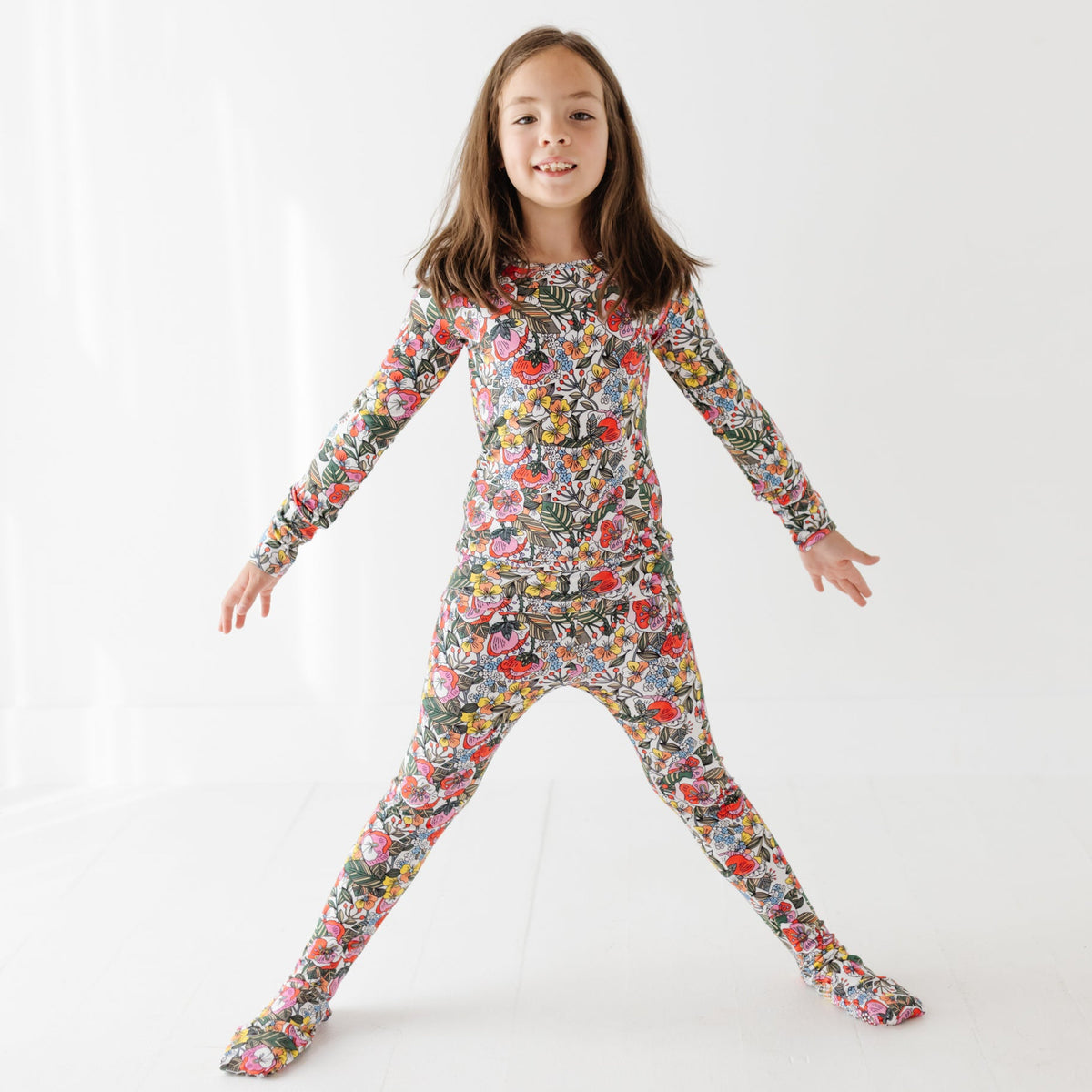 A smiling girl stands with her arms out on a white floor wearing a floral pajama set. 