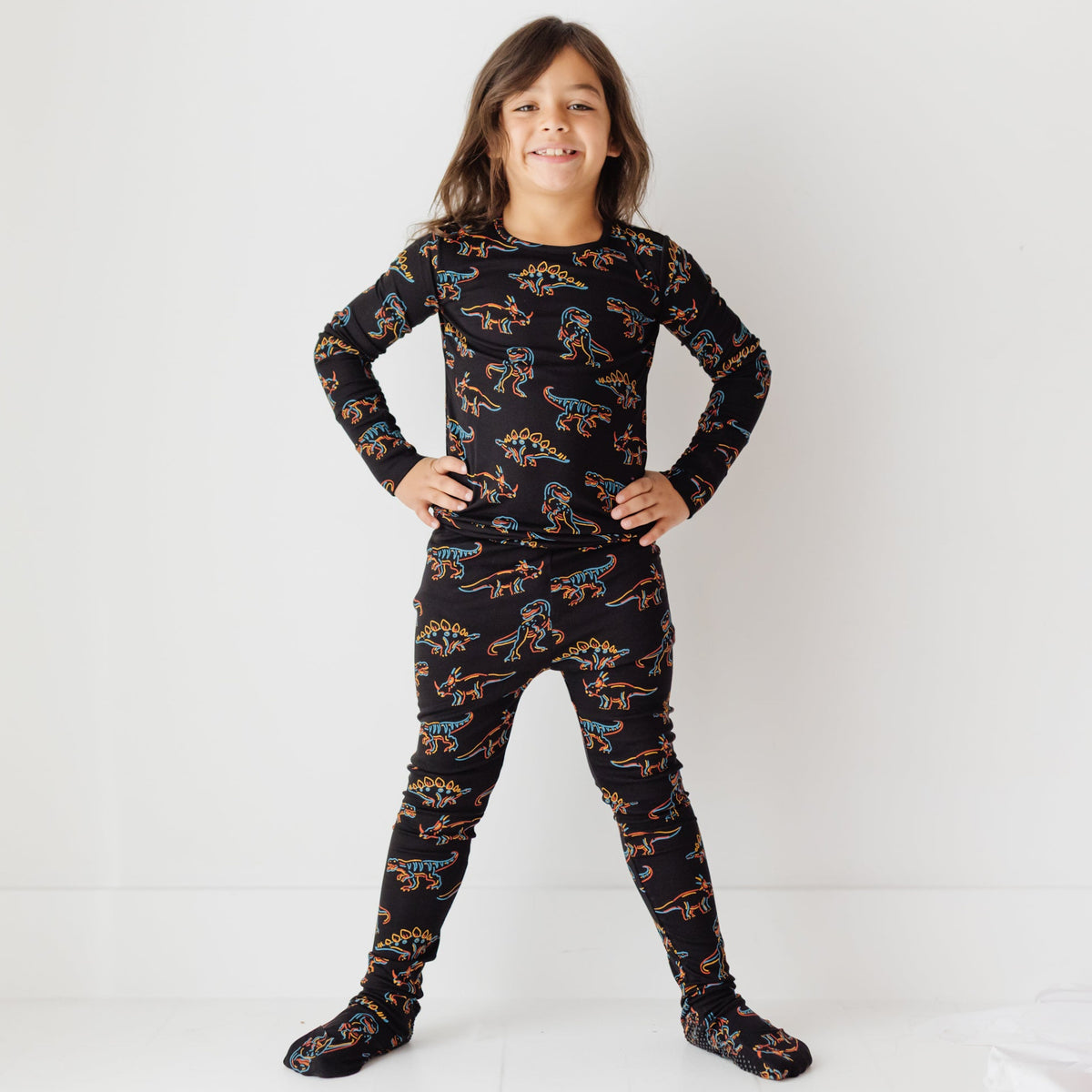 A smiling kid stands with hands on hips on a white floor, wearing a black pajama set with a neon dinosaur-themed print.