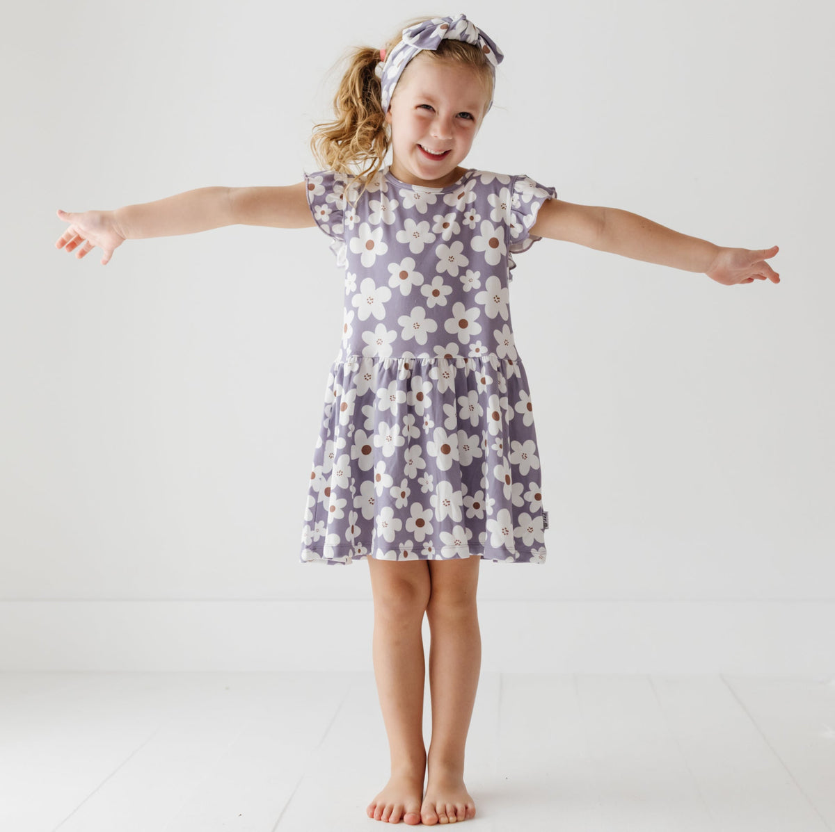 A smiling girl stands with her arms out on a white floor. She is wearing a lavender dress with white daisies on it. 