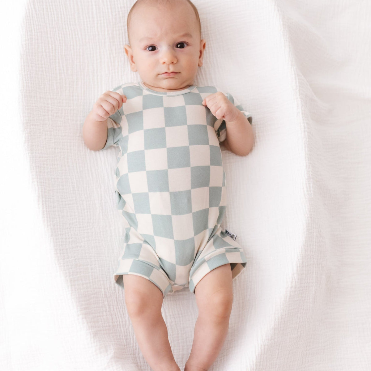 Baby wearing a checkered onesie lying on a white blanket