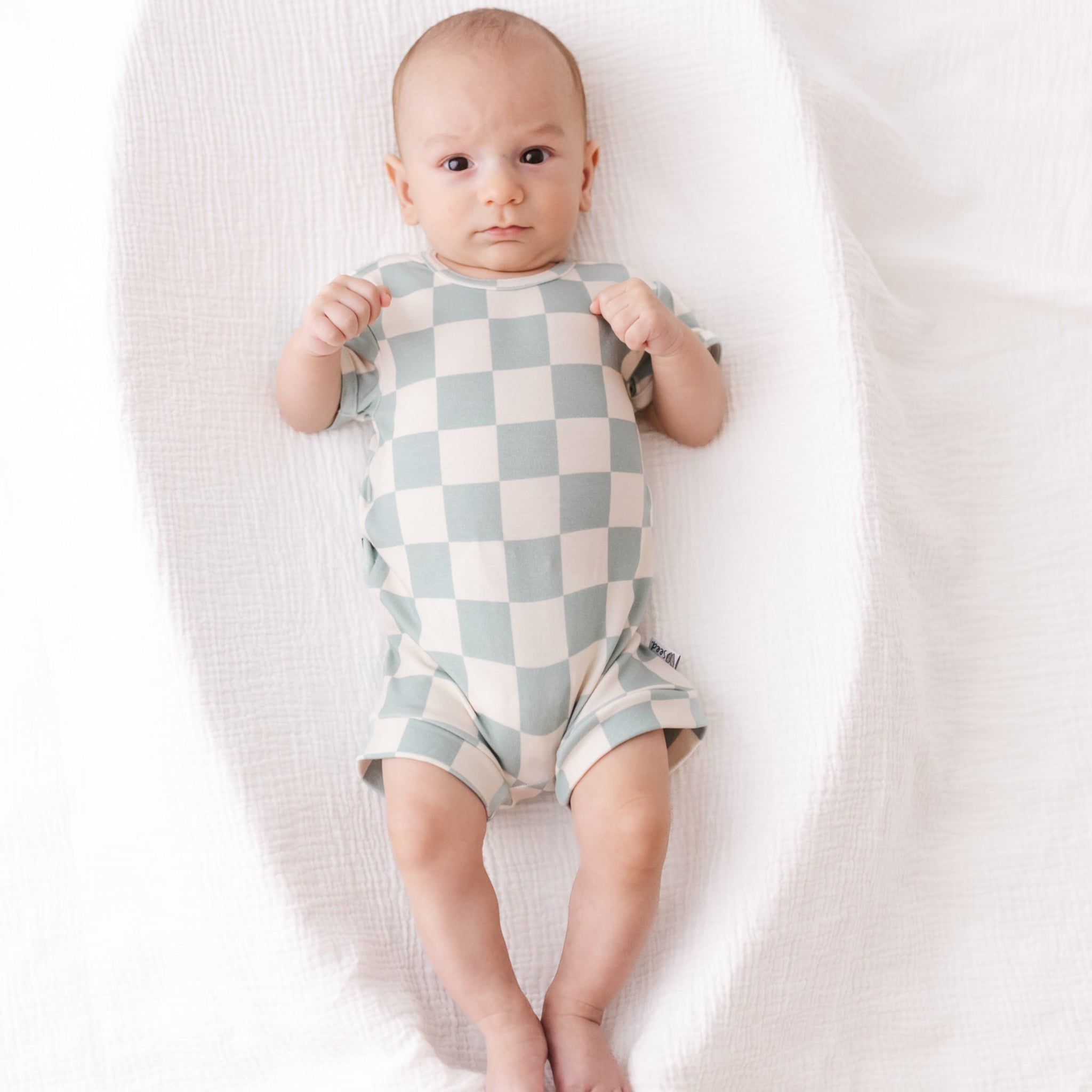 Baby wearing a checkered onesie lying on a white blanket