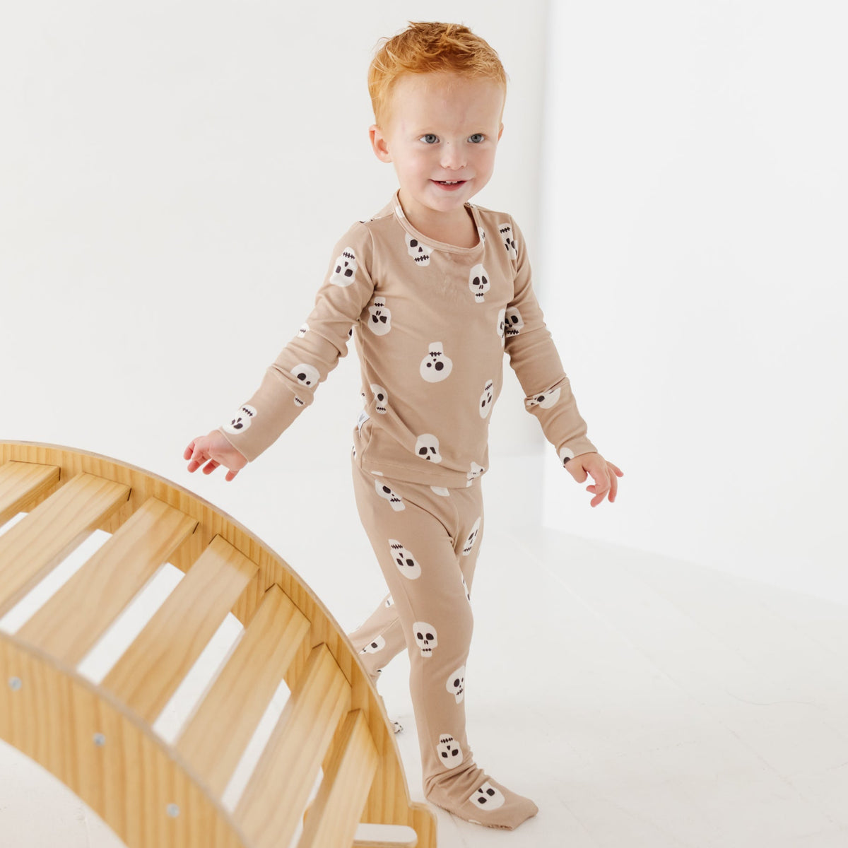 A smiling kid stand next to a wooden arch on a white floor wearing a tan pajama set with skulls on it. 