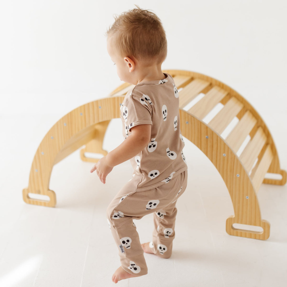 A kid stands next to a wooden arch on a white floor wearing a tan short-sleeved romper with skulls on it. 