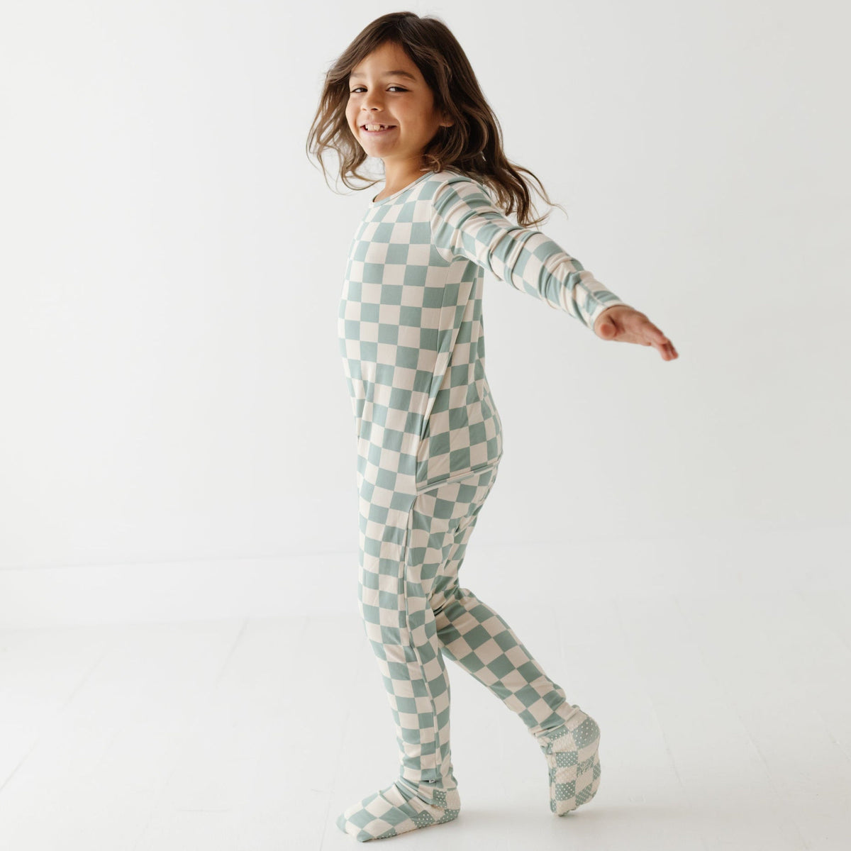 A kid stands on white floor wearing turquoise and white checked romper. 