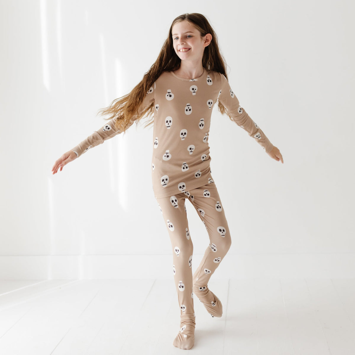 A smiling girl stands with her arms out on a white floor. She is wearing a tan pajama set with skulls on it. 
