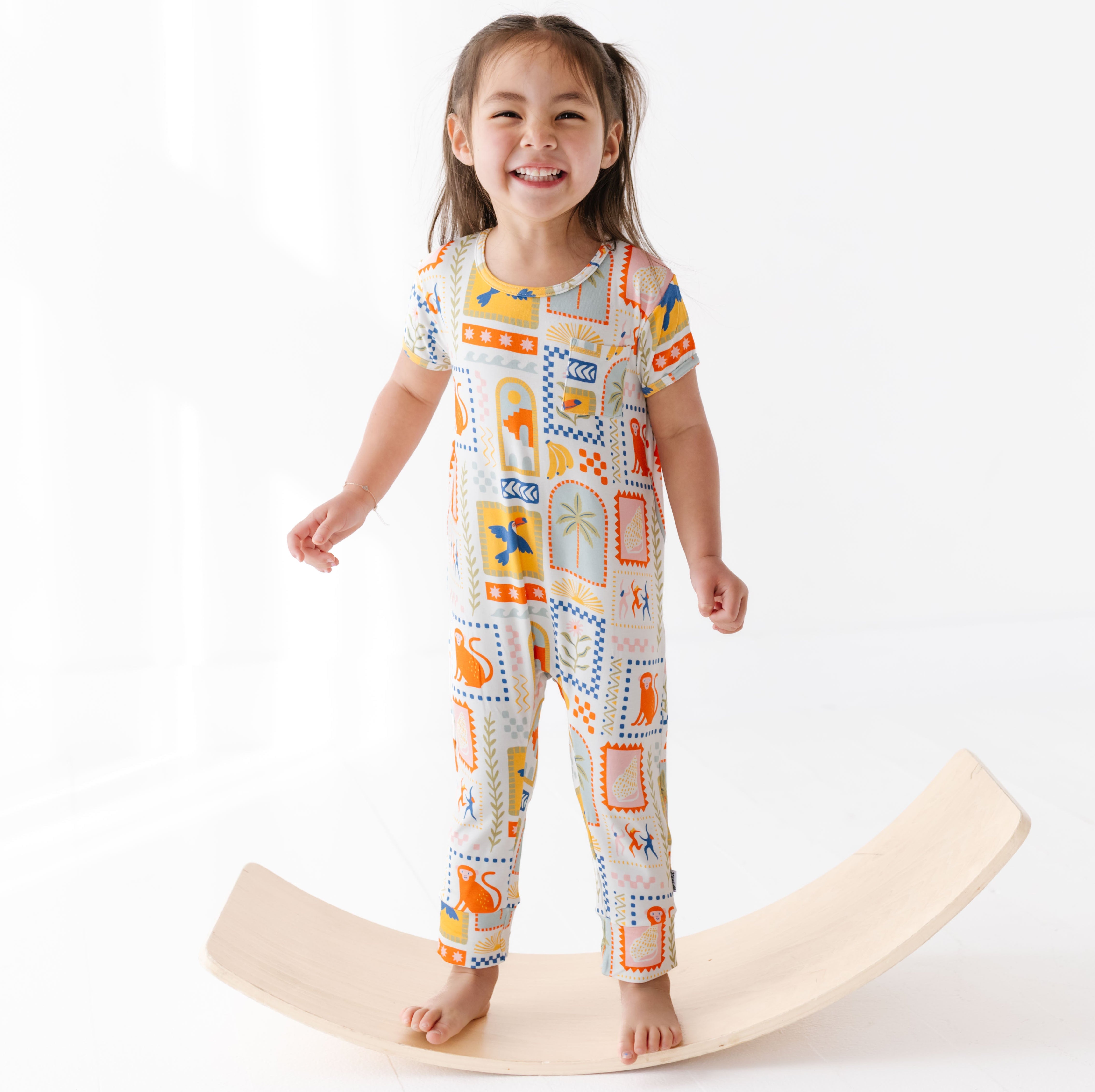 Child wearing a colorful patterned onesie standing on a wooden balance board against a white background