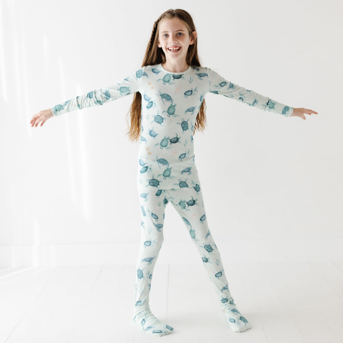 A kid stands on a white floor wearing a light green pajama set with turtles on it.