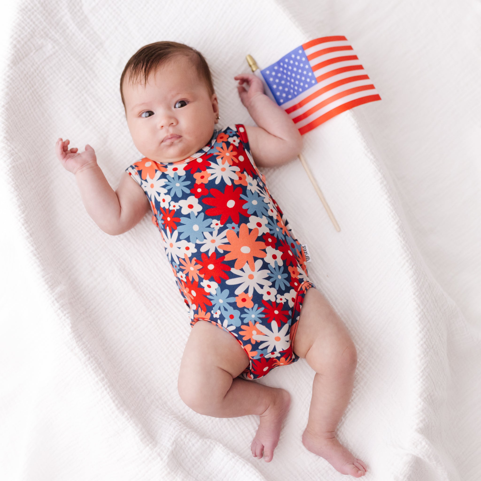 A baby lying on a white textured blanket, wearing a sleeveless red, white, and blue floral bodysuit and holding a small American flag.