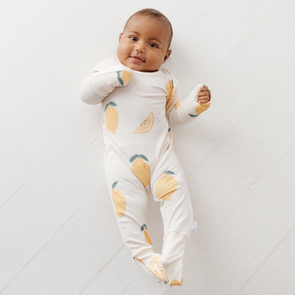 A baby lays on white floor wearing a cream color romper with lemons on it. 