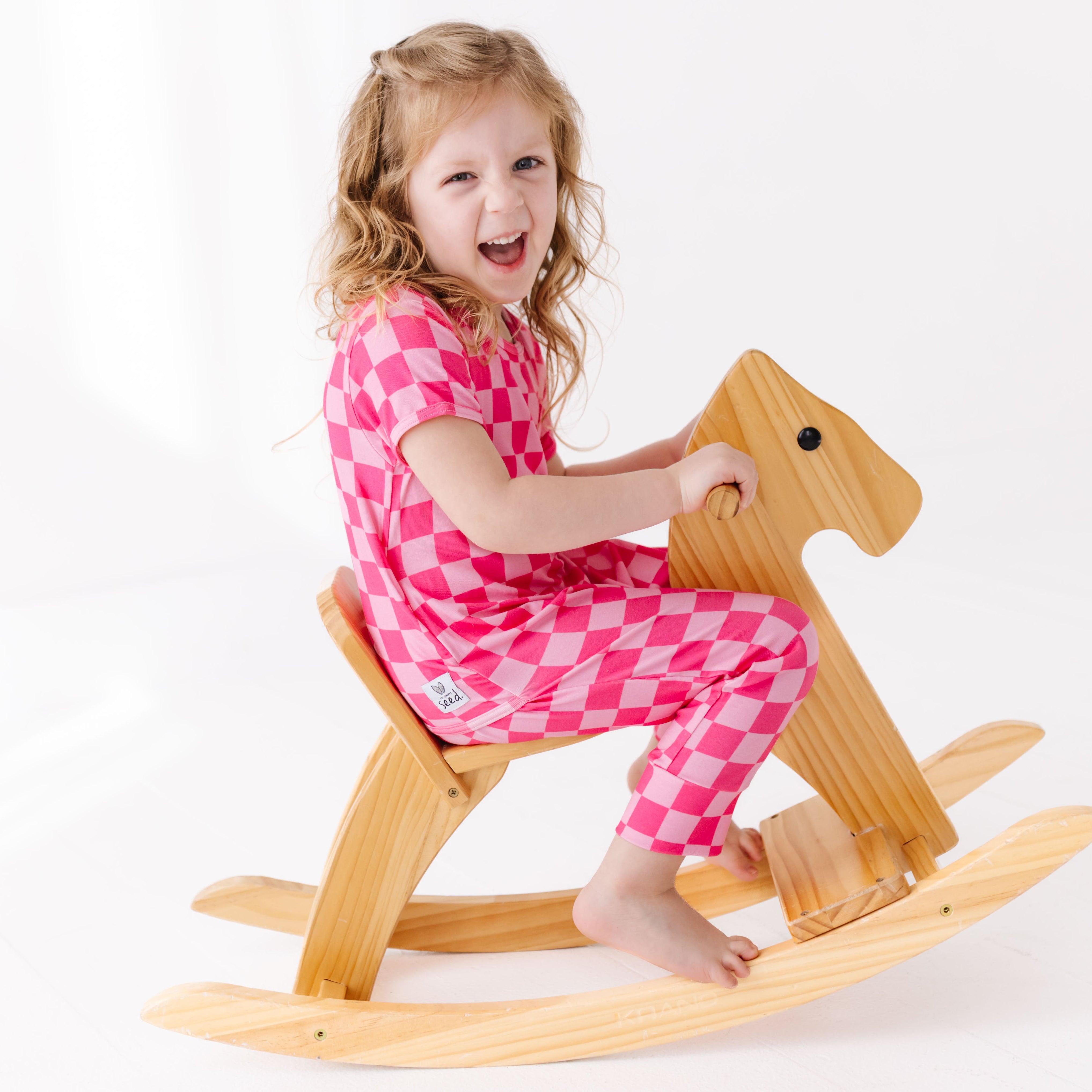 Child in pink checkered outfit riding a wooden rocking horse on a white background