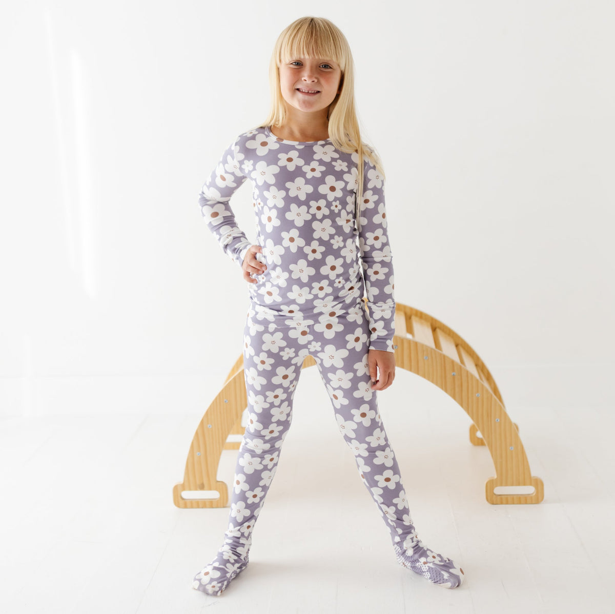 A smiling kid stands on a white floor with a wooden arch in the background. She is wearing a lavender outfit with white daisies on it. 