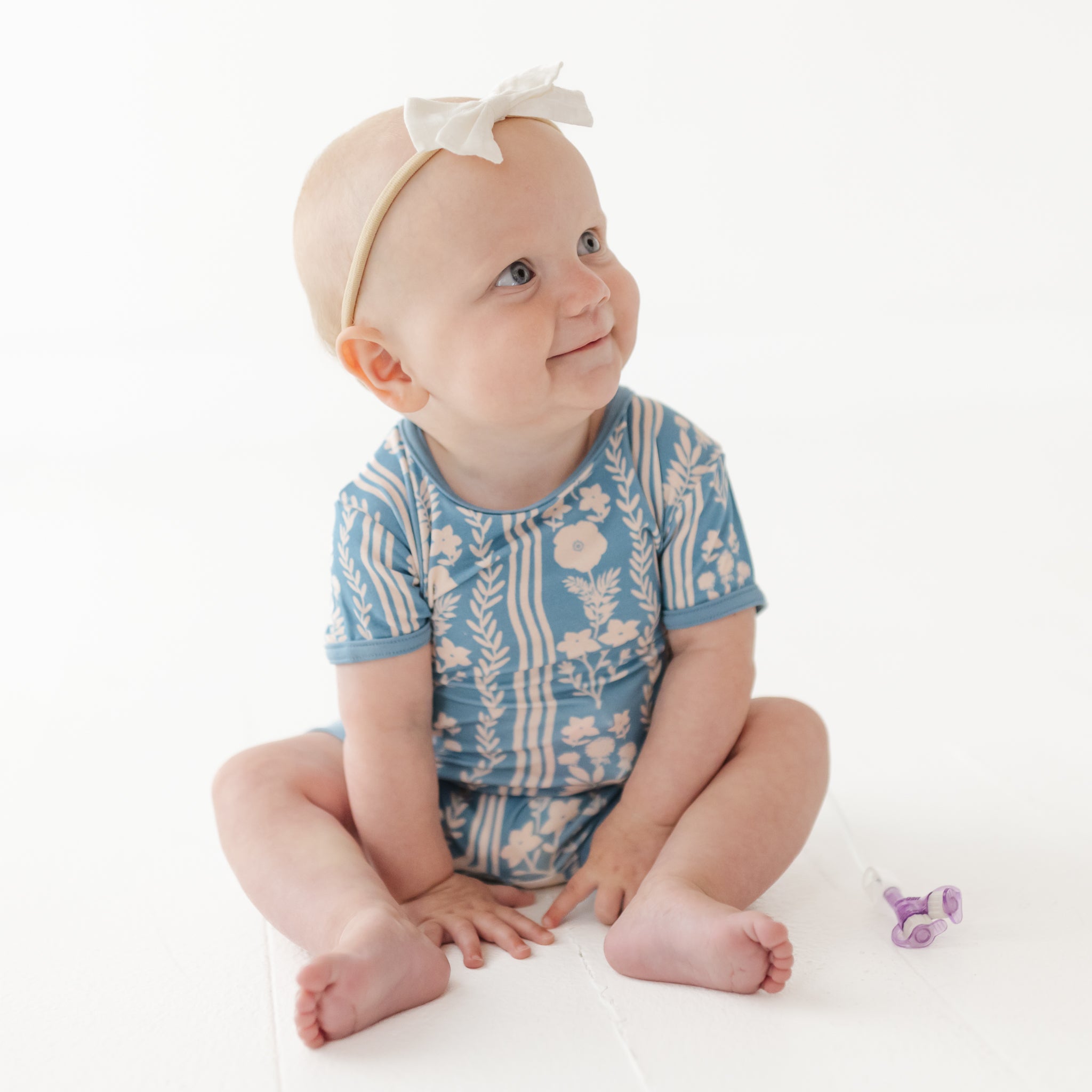 Baby wearing a blue floral onesie sitting on a white surface.