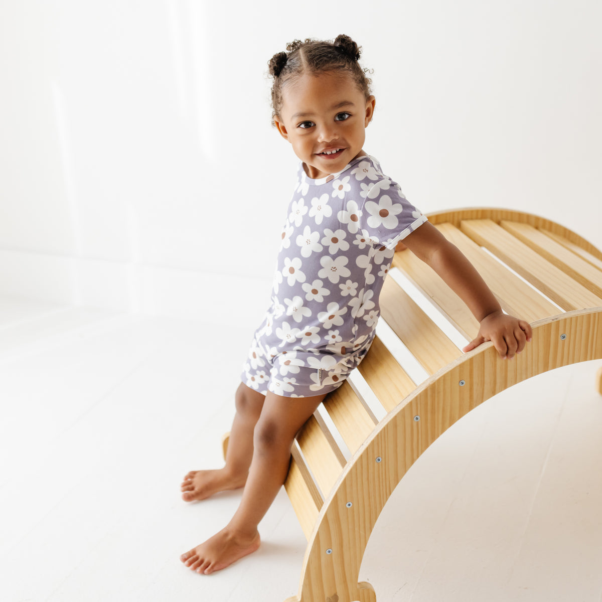 A smiling girls leans on a wooden arch on a white floor. She is wearing a lavender short-sleeved romper with white daisies on it. 