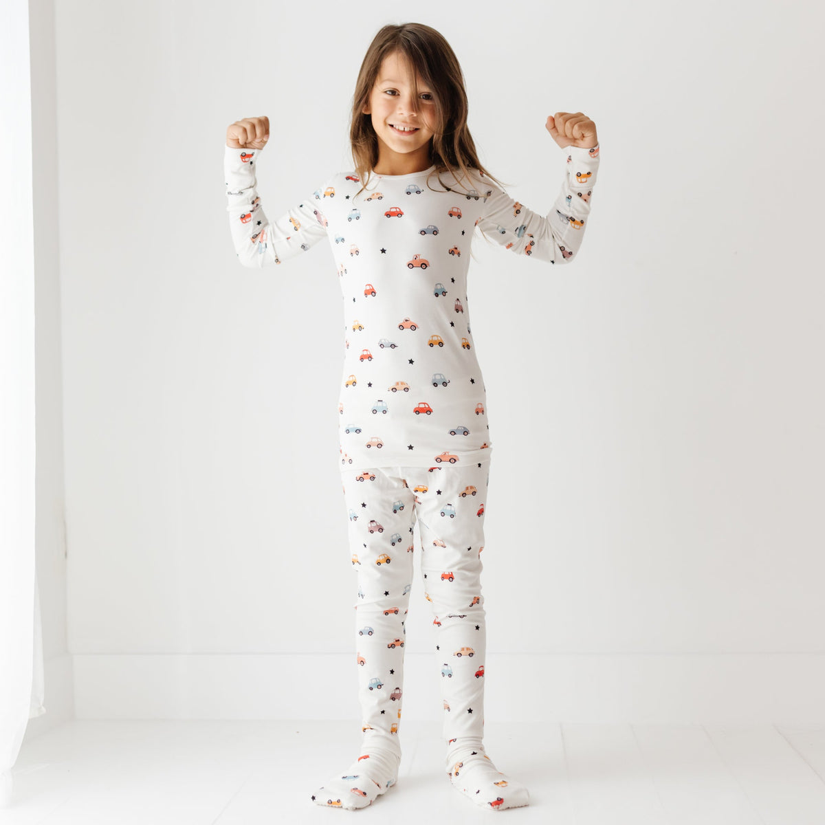 A smiling kid stands on a white floor, wearing a white pajama set with a car-themed print.