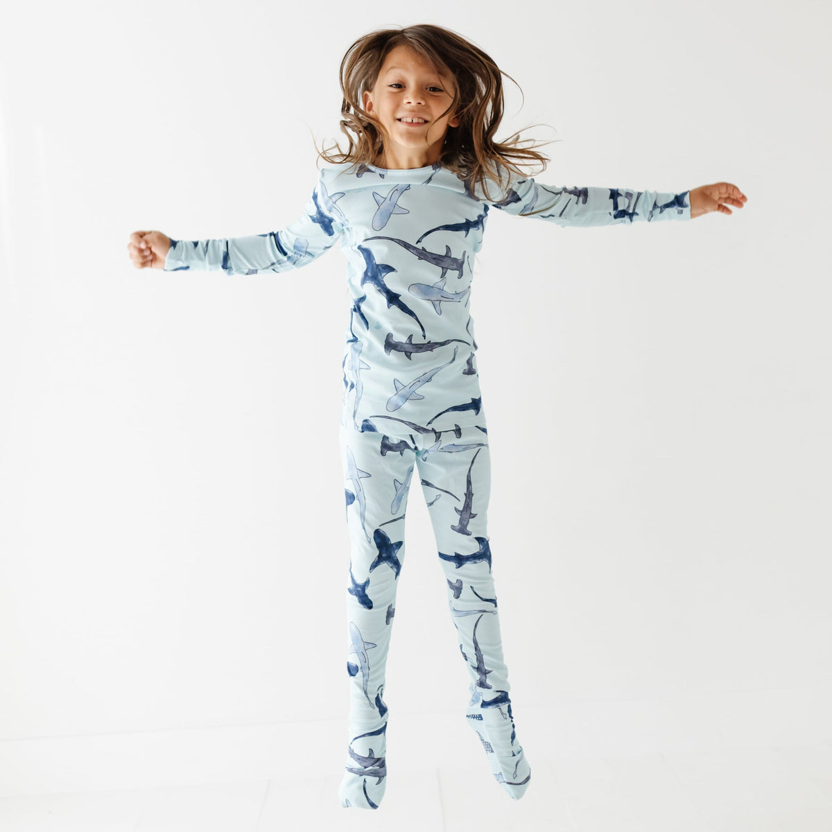A kid jumps in the air wearing a light blue pajama set with different shades of blue sharks on it. 