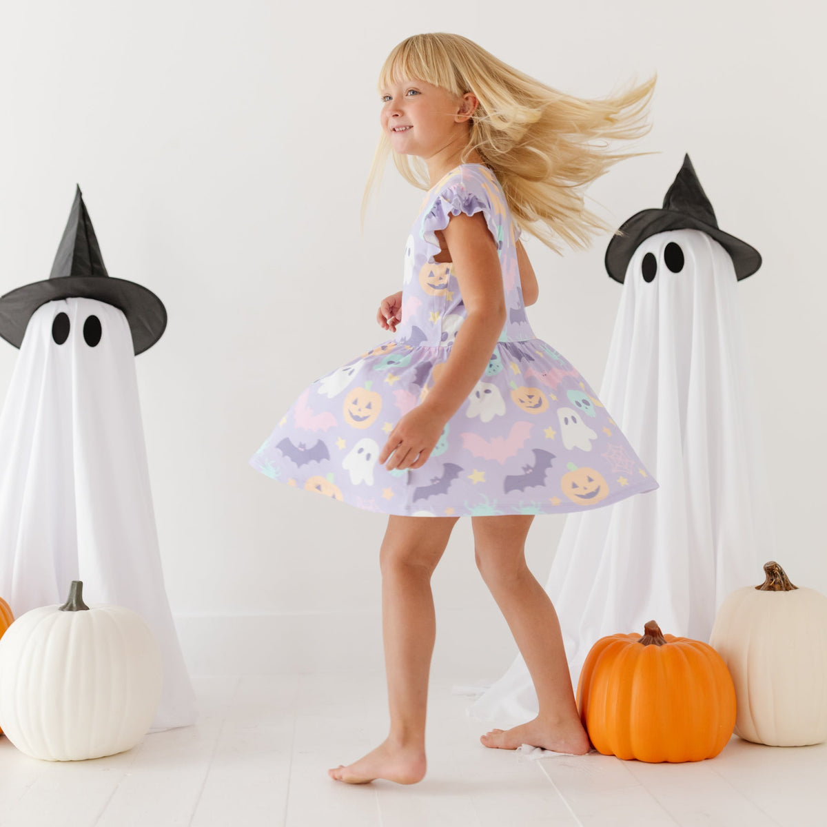 A girl spins on a white floor with pumpkins and ghosts in the background. She is wearing a lavender dress and shorts with a Halloween-themed print.