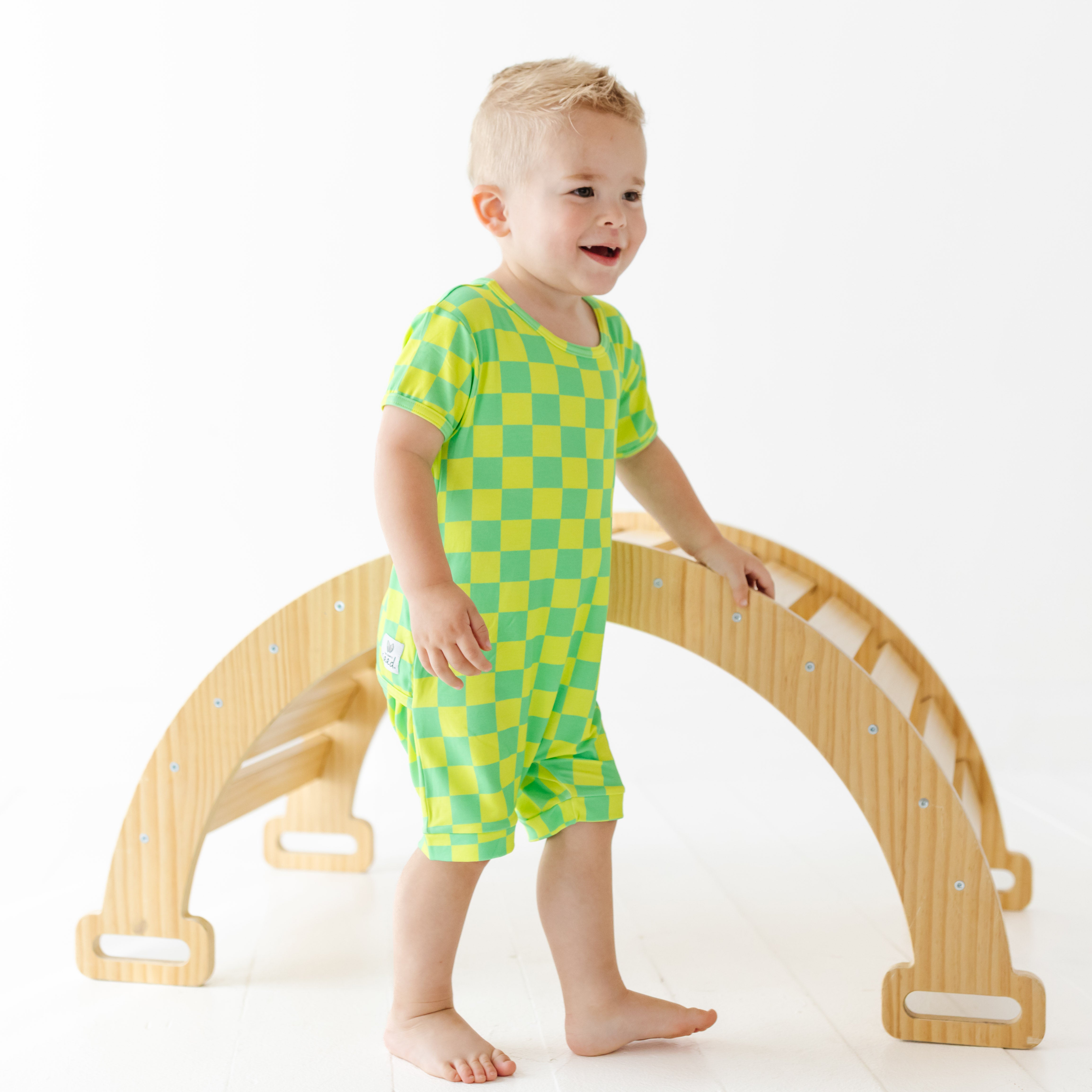 Child in green checkered outfit standing next to a wooden arch toy on a white background