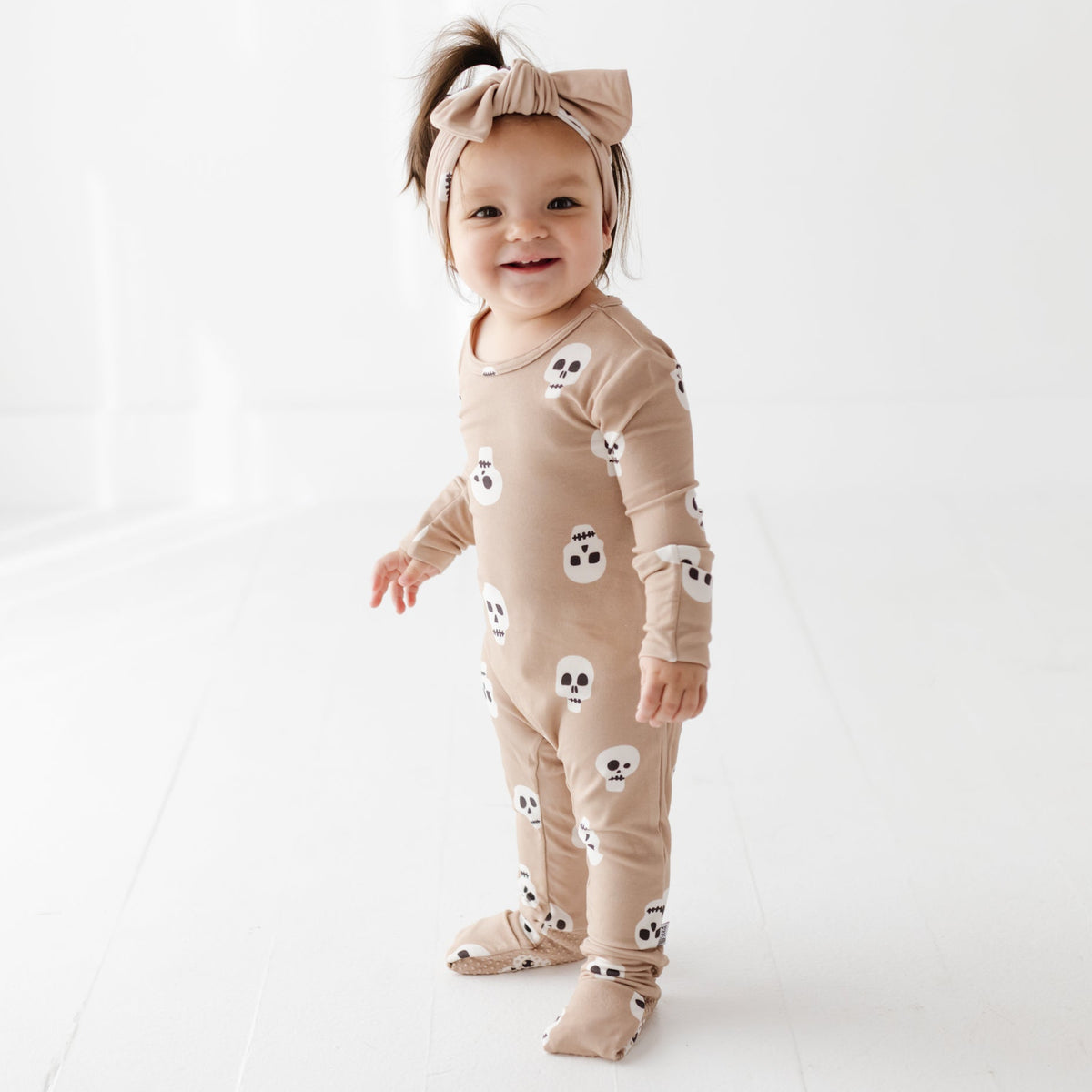 A smiling kid stand next to a wooden arch on a white floor wearing a tan romper with skulls on it. 