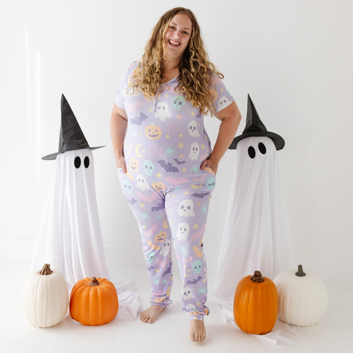 A smiling adult stands on white floor wearing lavender pajama set with Halloween design on it. 