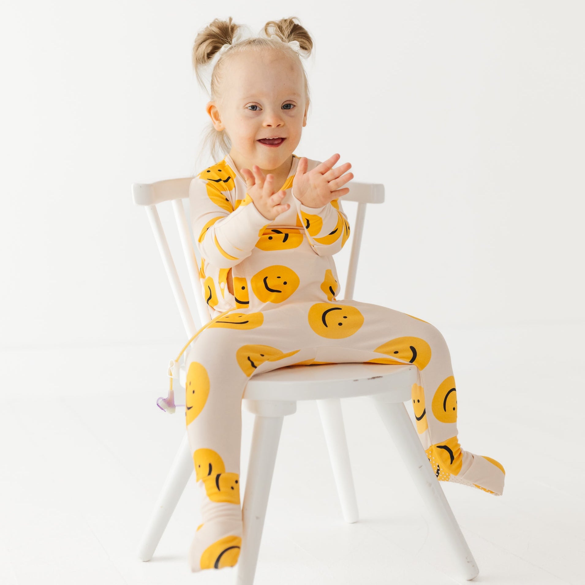 Child wearing a onesie with smiley face patterns sitting on a chair against a white background
