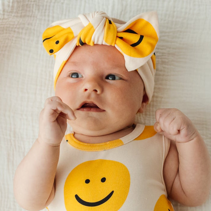 A baby lying on a white muslin blanket, wearing a cream bodysuit with yellow smiley face prints and a matching yellow smiley headband.