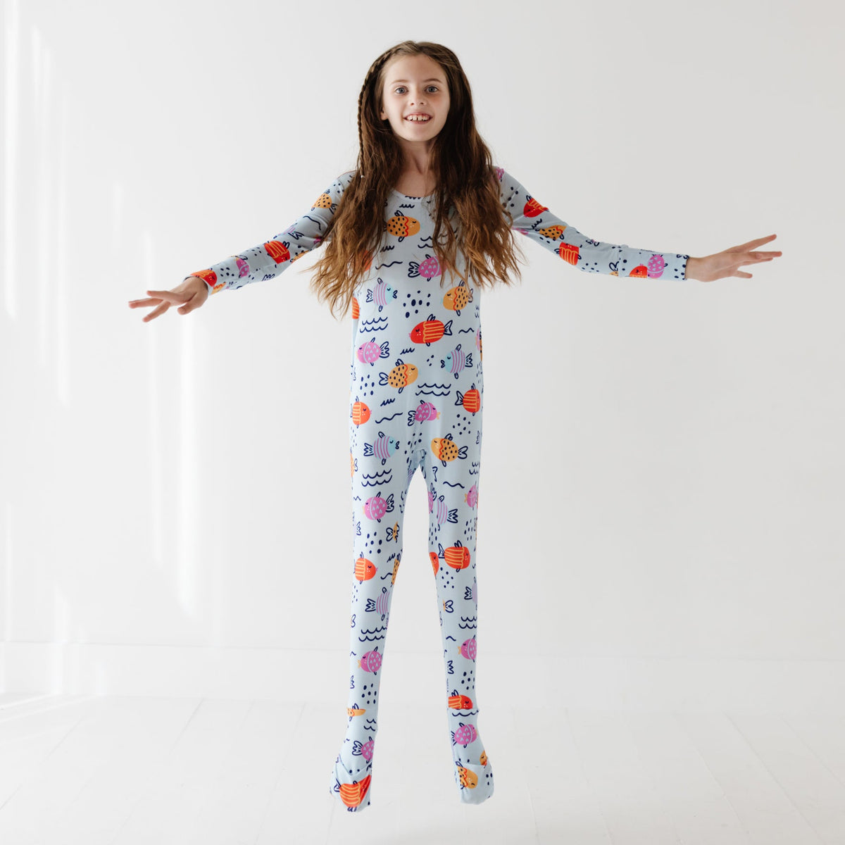 A kid jumps off of white floor wearing a light blue romper with colorful fish on it. 