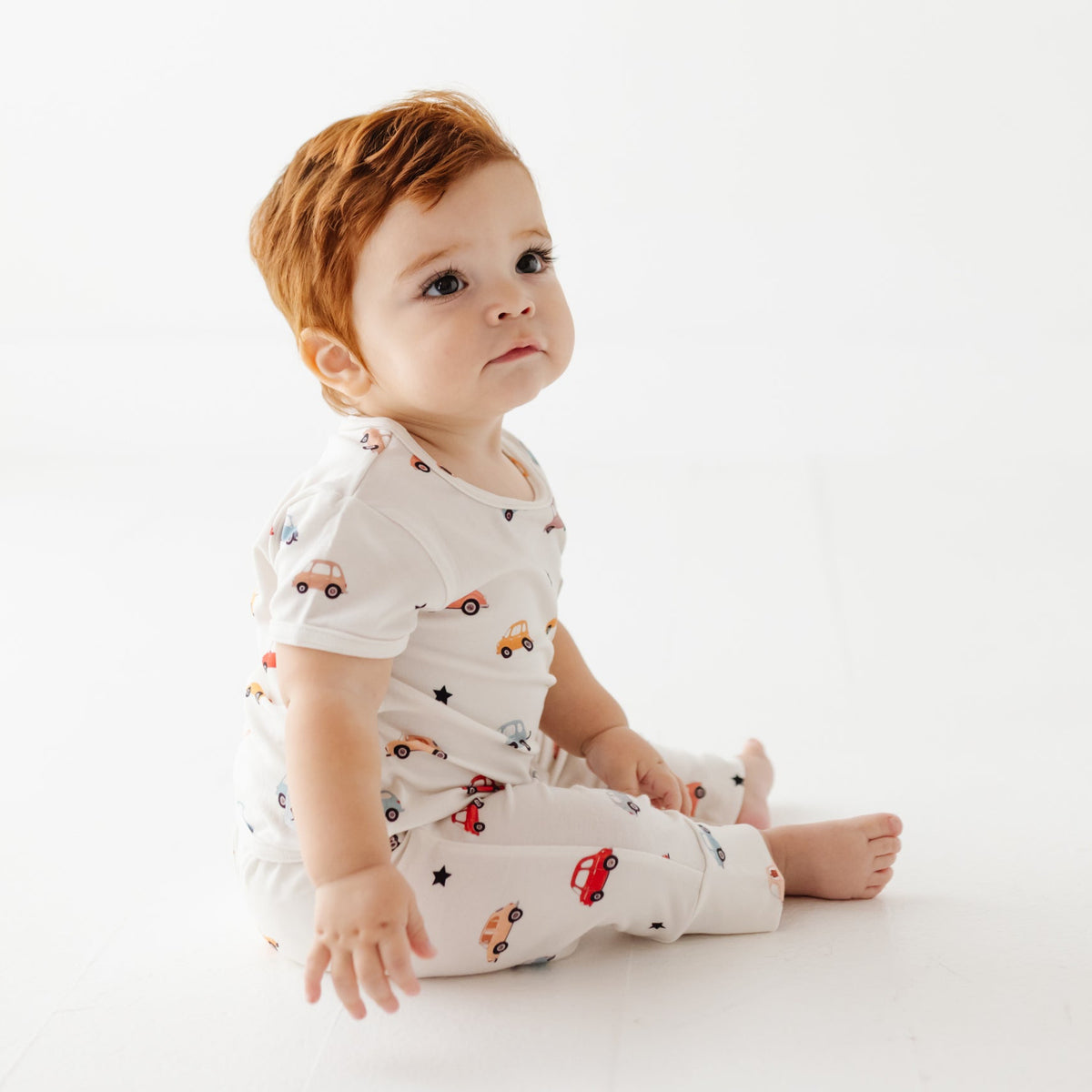 A baby sits on white floor wearing white romper with car print. 