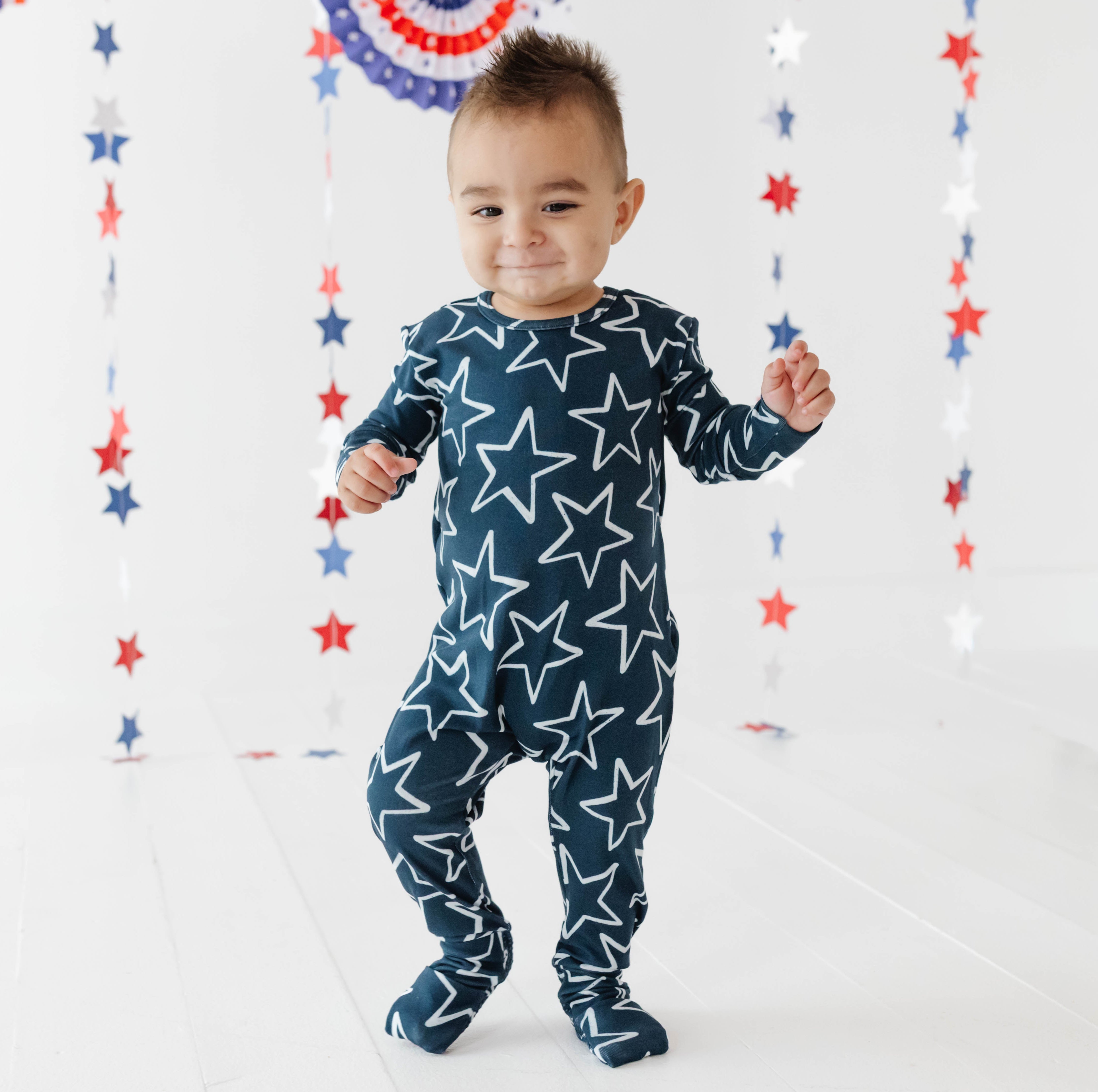 Child wearing a navy blue onesie with white star patterns against a decorative background with red, white, and blue stars.