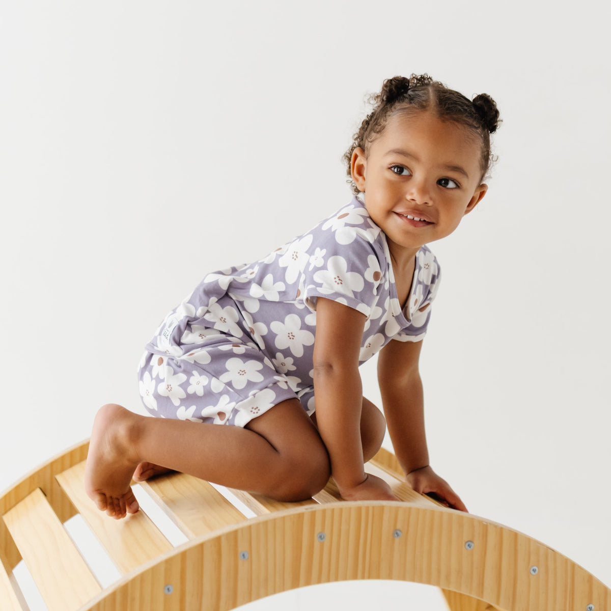 A smiling girls climbs on a wooden arch on a white floor. She is wearing a lavender short-sleeved romper with white daisies on it. 