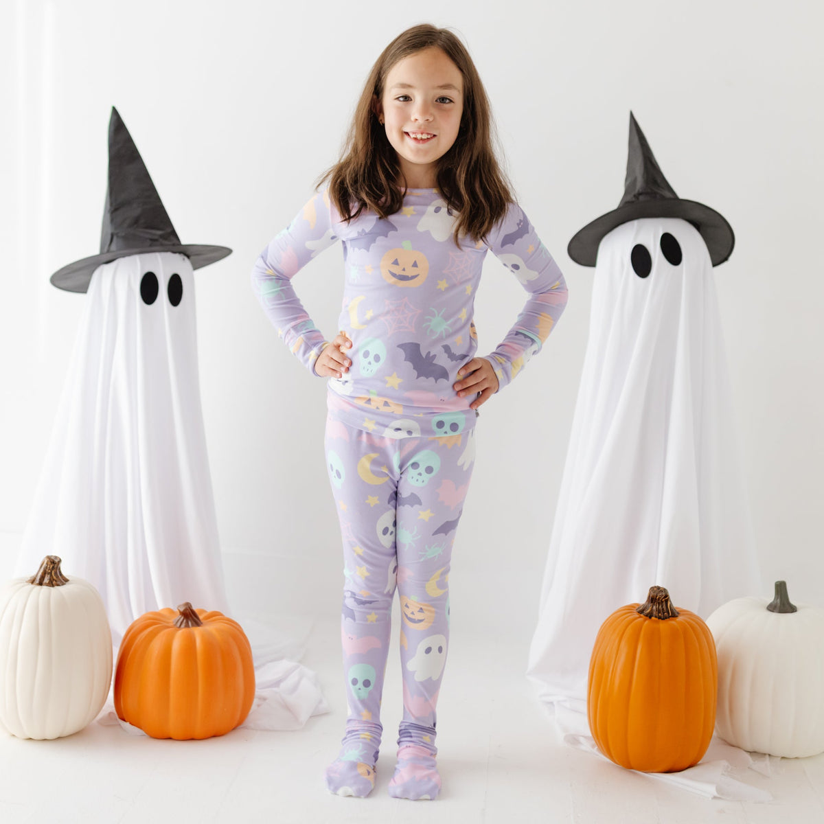 A smiling kid stands on white floor wearing lavender pajama set with Halloween design on it. 