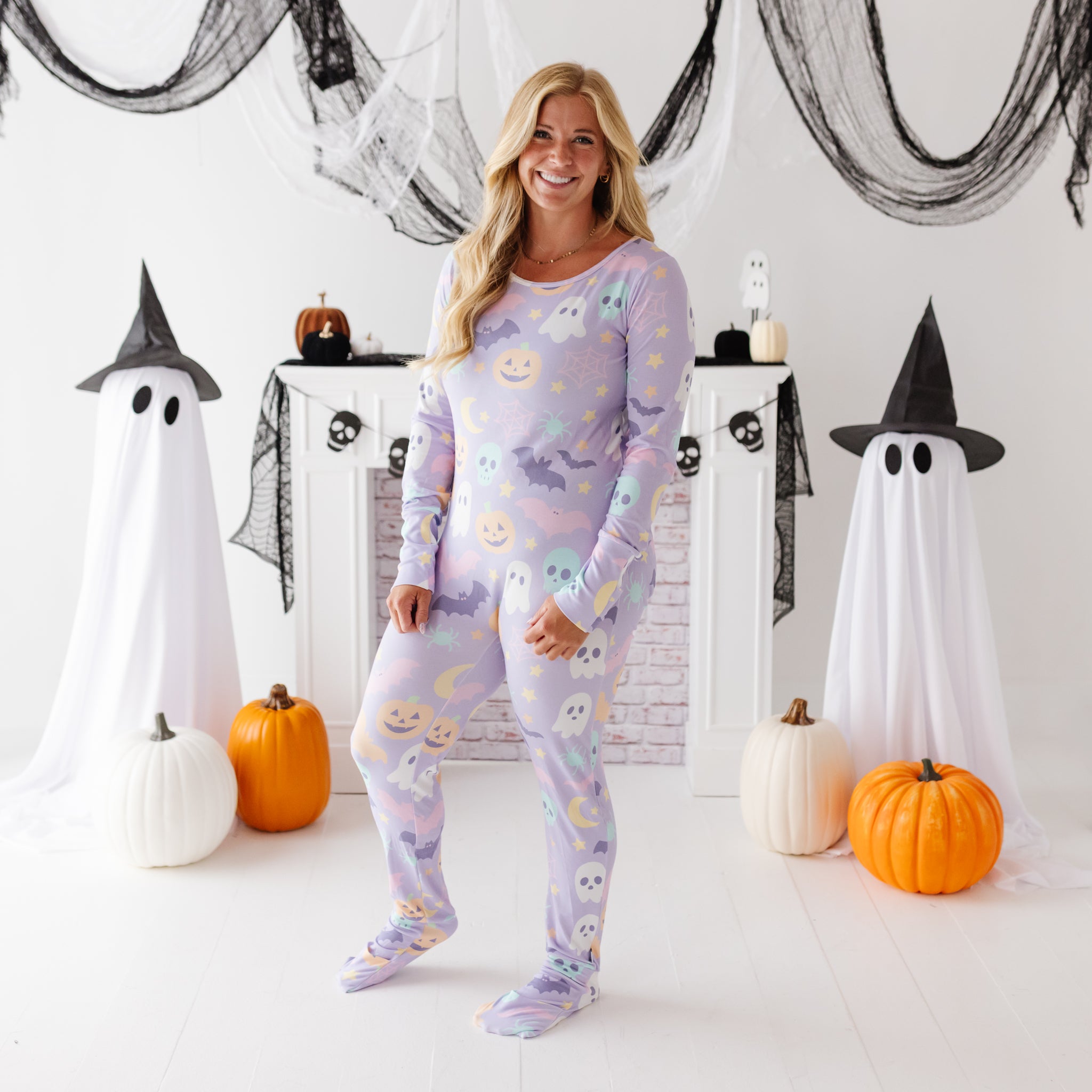 A woman stands in a Halloween-themed room wearing a light lavender long-sleeve pajama set with a cute ghost and moon pattern. The room is decorated with white ghost figures draped in black sheets, orange pumpkins, and black netting.