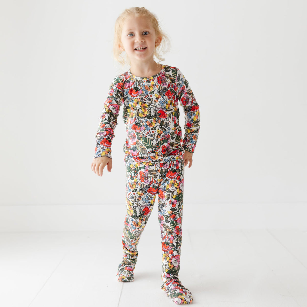 A smiling girl stands on a white floor wearing a floral pajama set. 