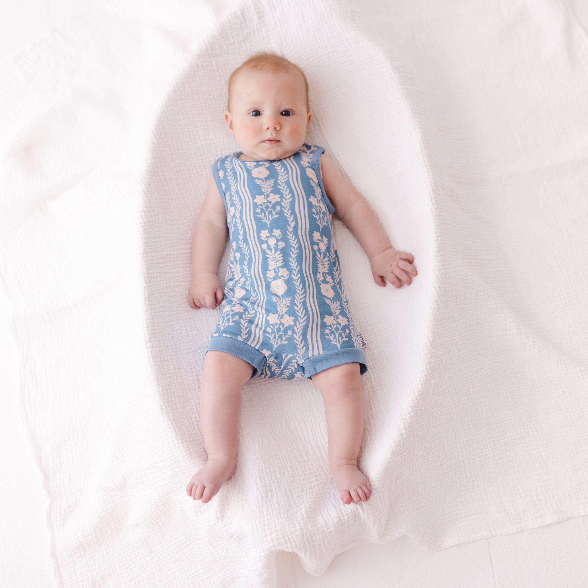 A baby lying on a white textured blanket, wearing a sleeveless light blue romper with a white floral pattern.
