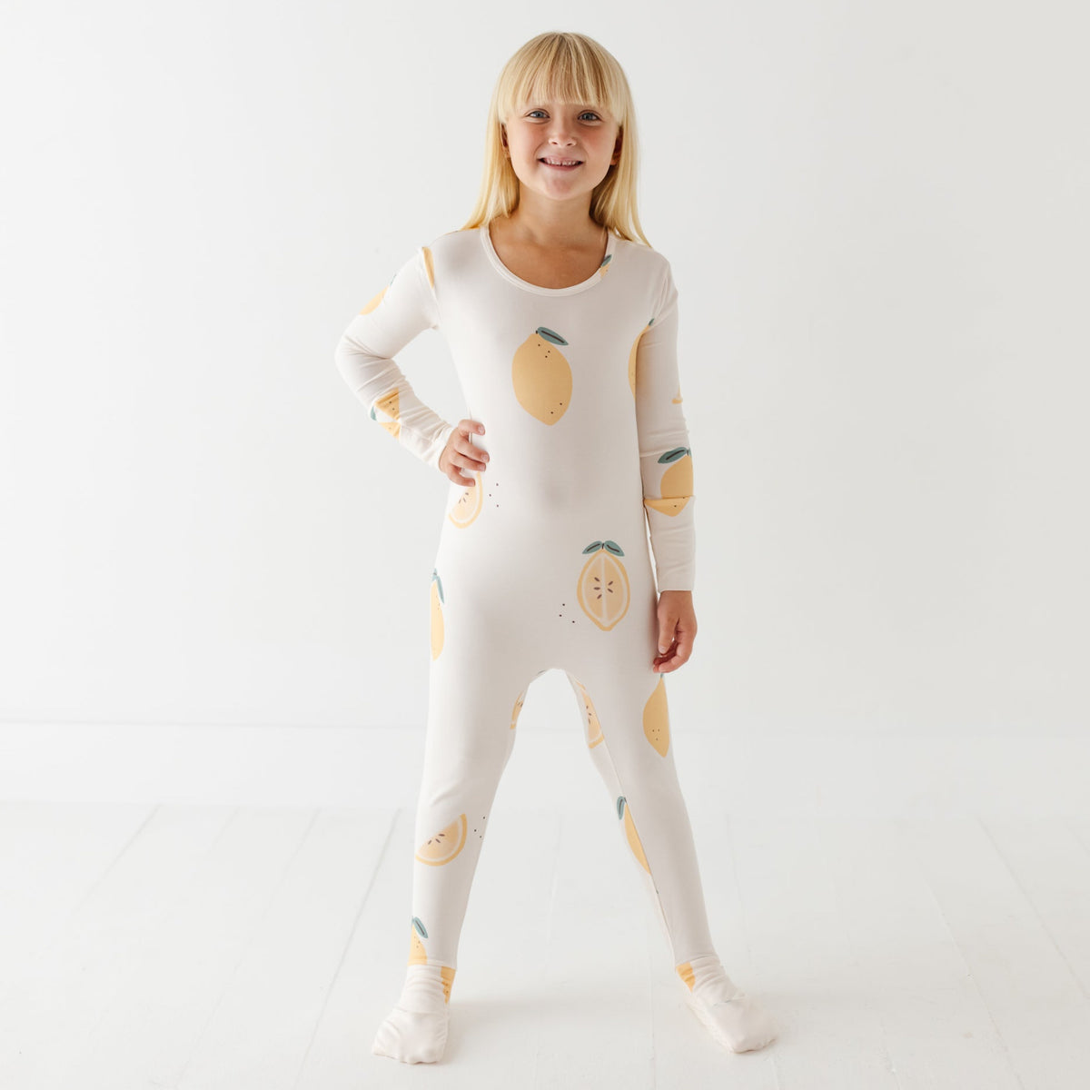 A smiling girl stand on white floor wearing a cream rompers with lemons on it. 