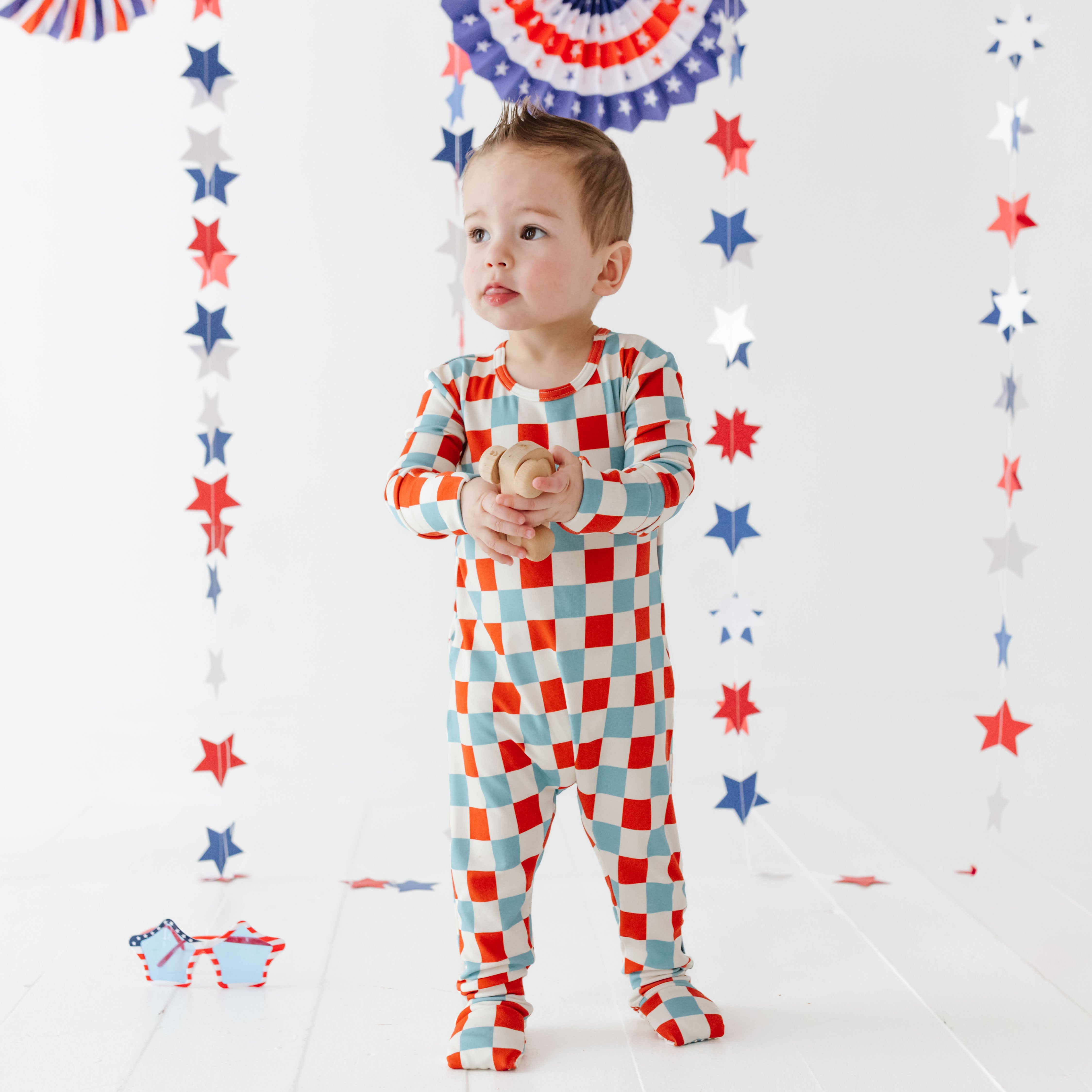 Child wearing a red, white, and blue checkered outfit with decorative stars and stripes in the background.