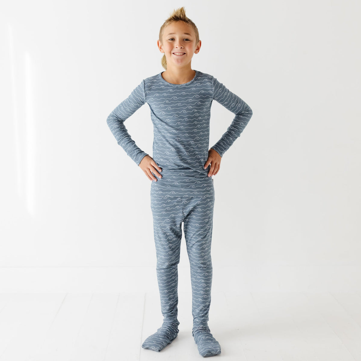 A smiling kid stands with his hands on his hips on a white floor. They are wearing blue pajama set with waves in it. 