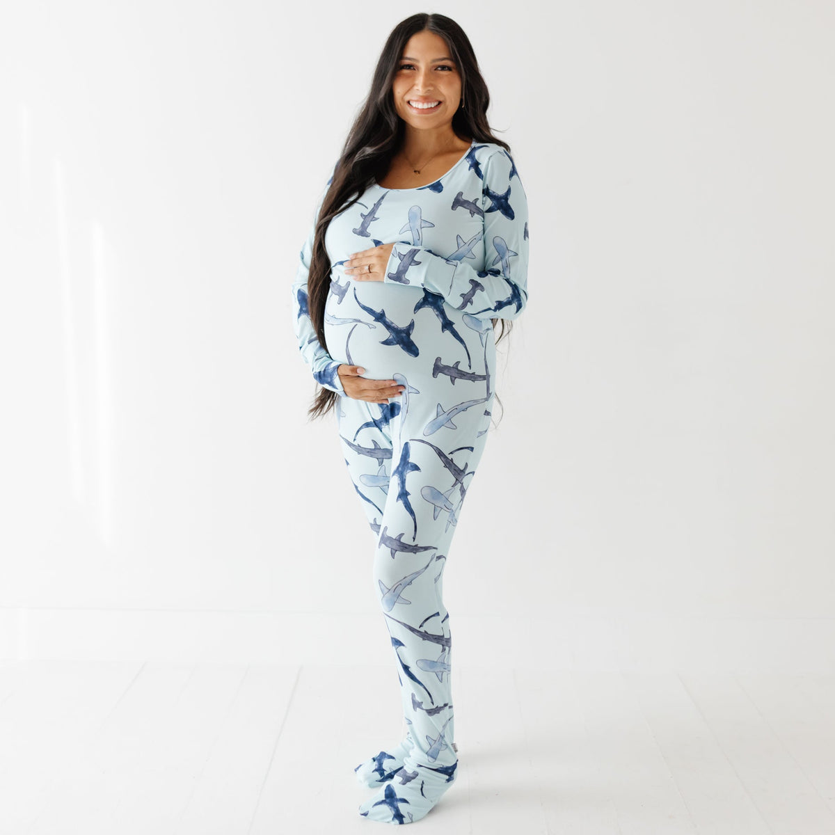 A smiling woman stands on a white door wearing a light blue romper with different shade of blue sharks on it. 