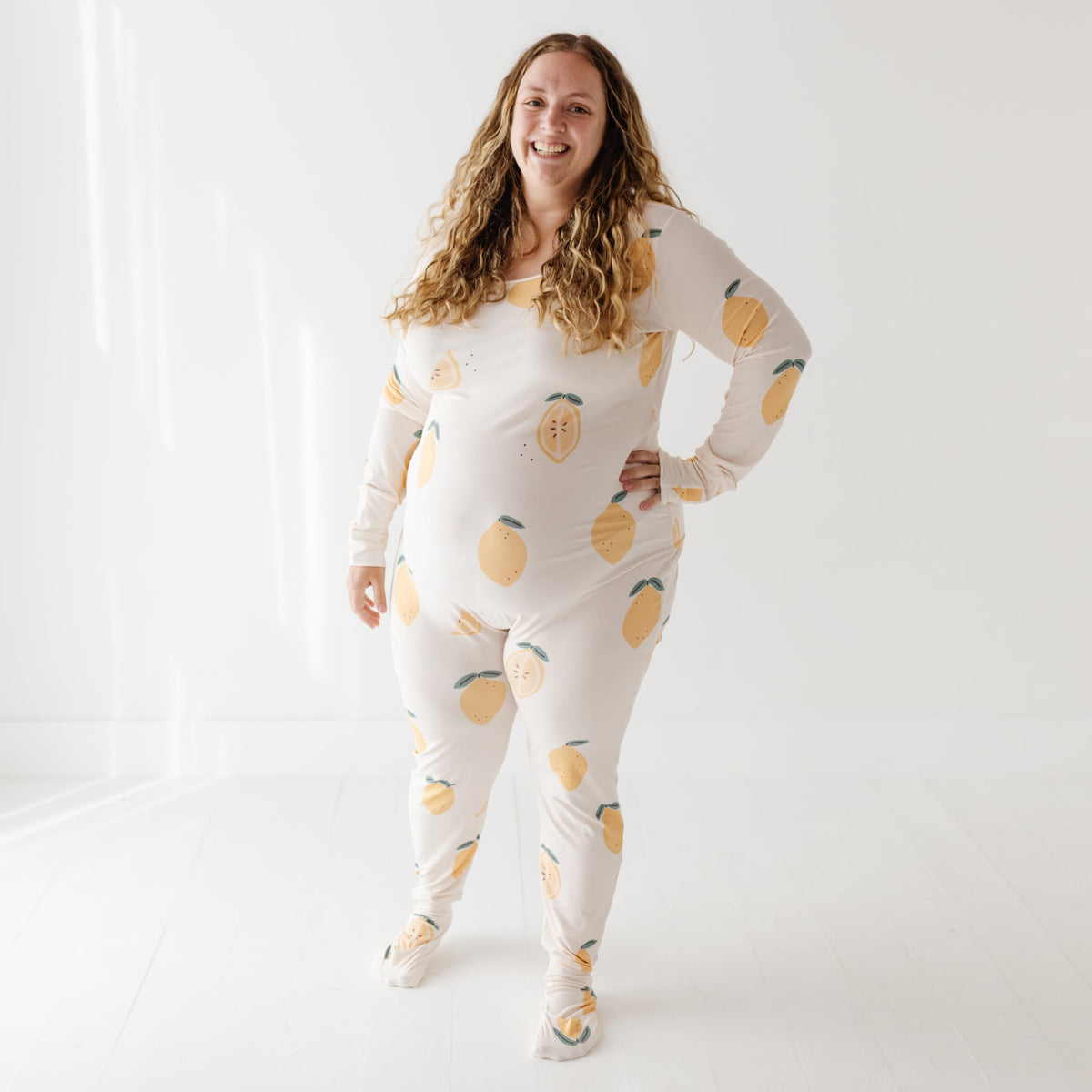 A woman stands on a white floor wearing a cream color romper with lemons on it. 