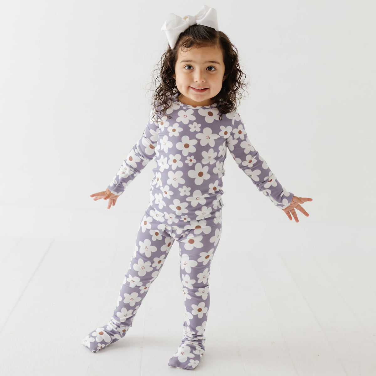 Girl stands on white floor wearing lavender pajama set with white daisies on it. 