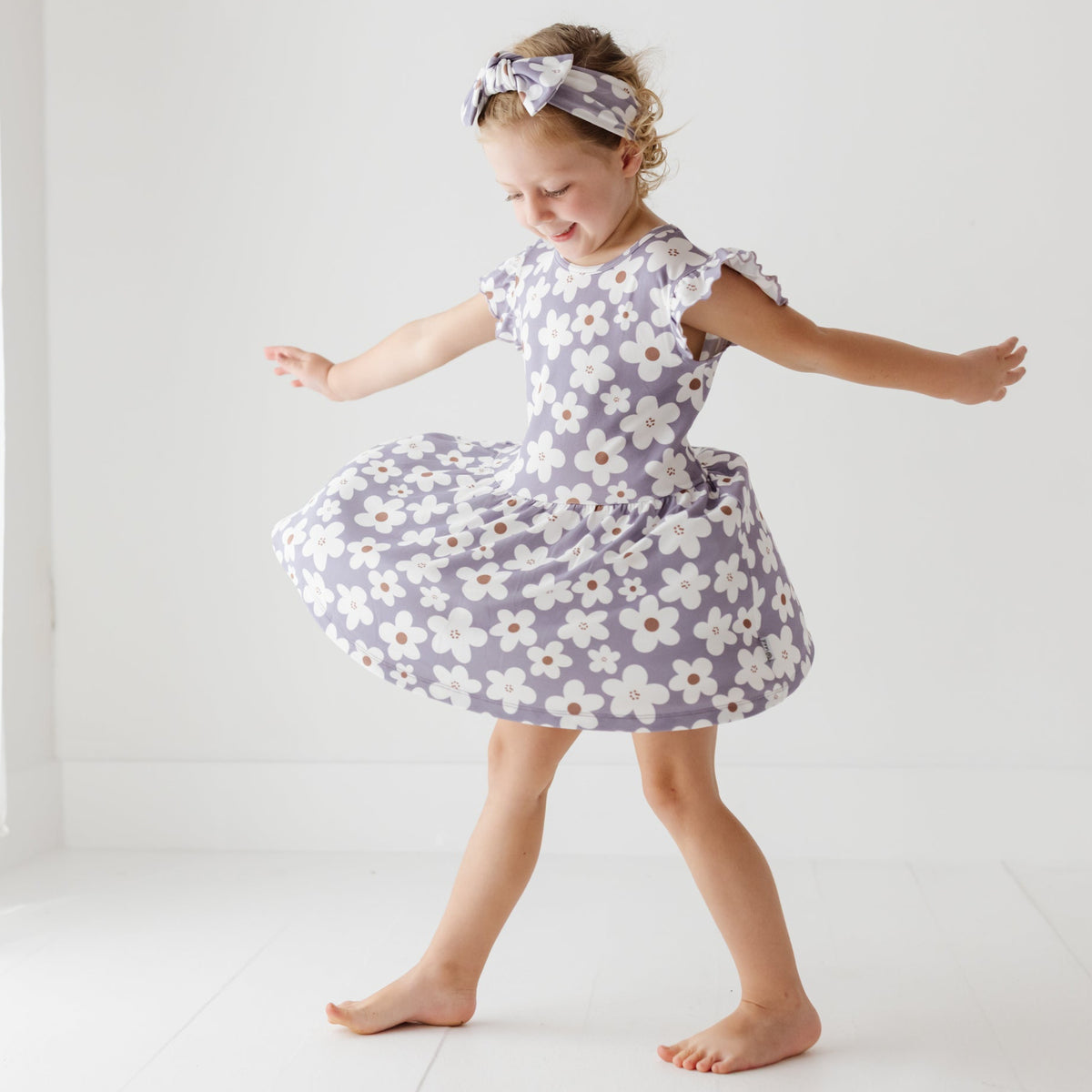 A smiling girl stands with her arms out on a white floor. She is wearing a lavender dress with white daisies on it. 