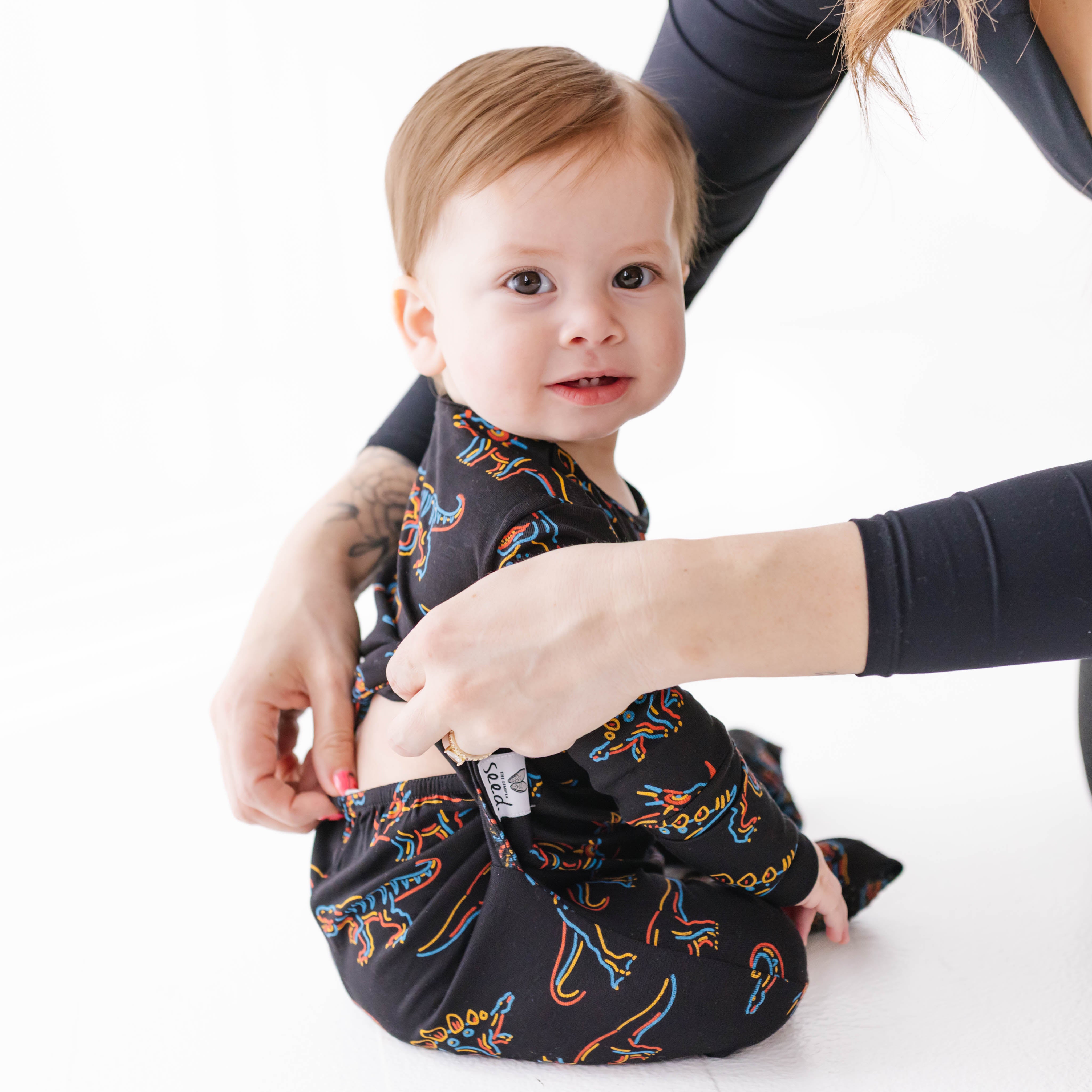 Baby wearing a dinosaur-patterned outfit with a person adjusting it on a white background